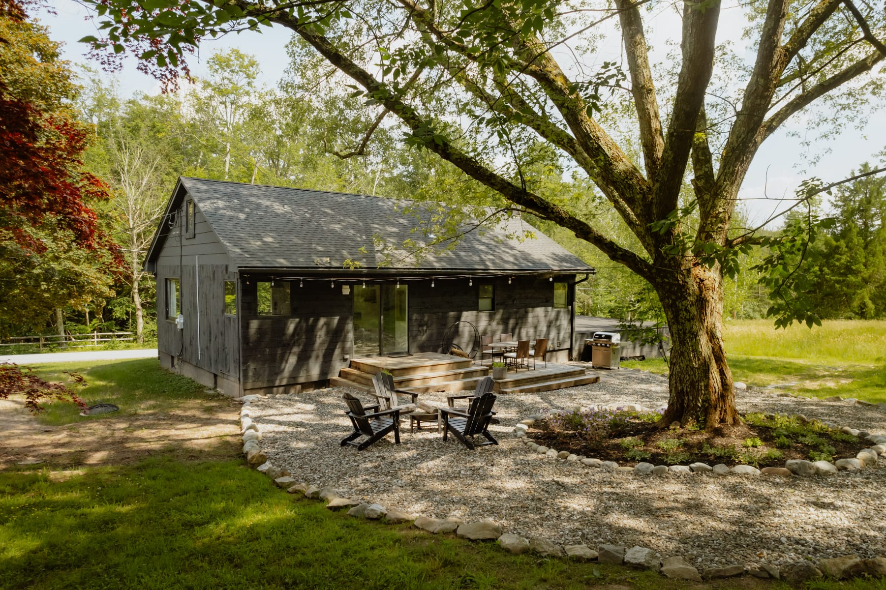 The rear view of a dark-sided cabin features a spacious wooden deck with outdoor seating and a BBQ grill, extending to a gravel patio area under a mature tree. A fire pit with Adirondack chairs is nestled within the gravel, surrounded by lush greenery.