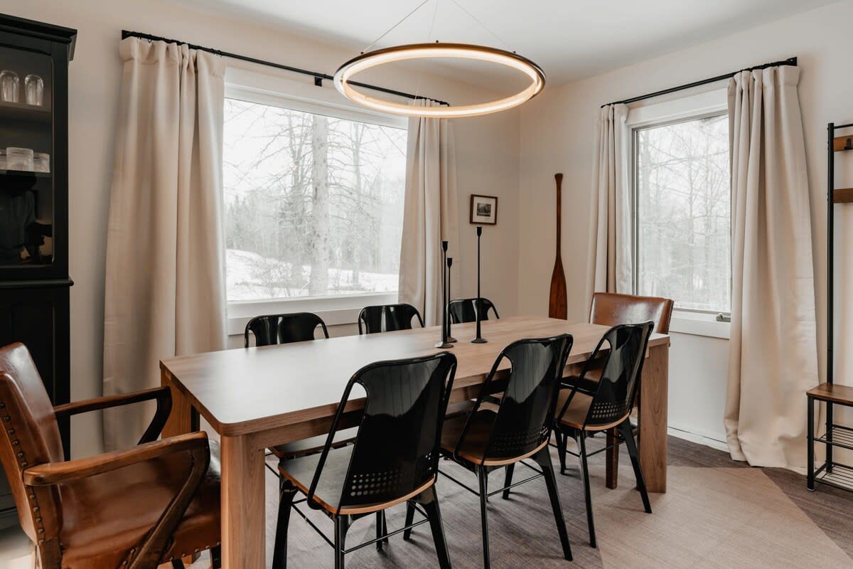 A modern dining room with a light wood rectangular table, black chairs, and two brown leather armchairs, illuminated by a large circular LED chandelier, with windows showing a snowy outdoor scene.