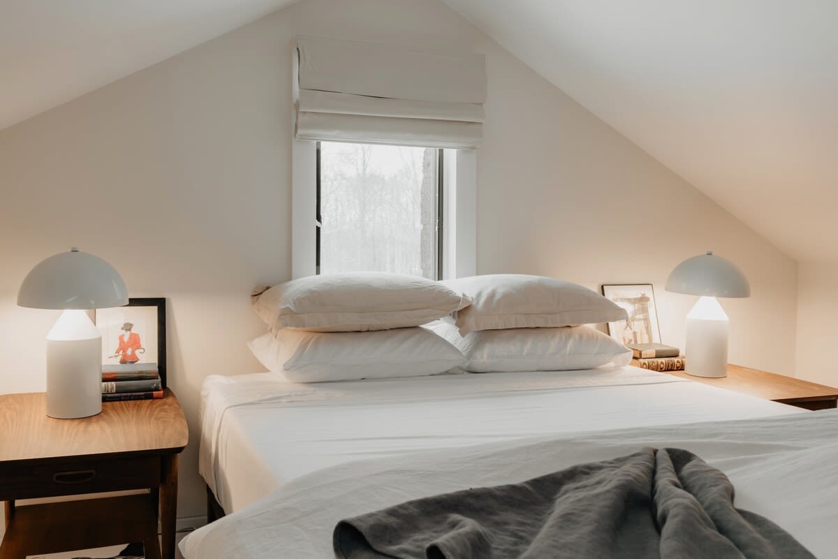 Close-up of the bed in the attic bedroom, showing crisp white bedding, multiple pillows, and a gray throw blanket. Two modern white table lamps flank the bed on simple wooden nightstands.