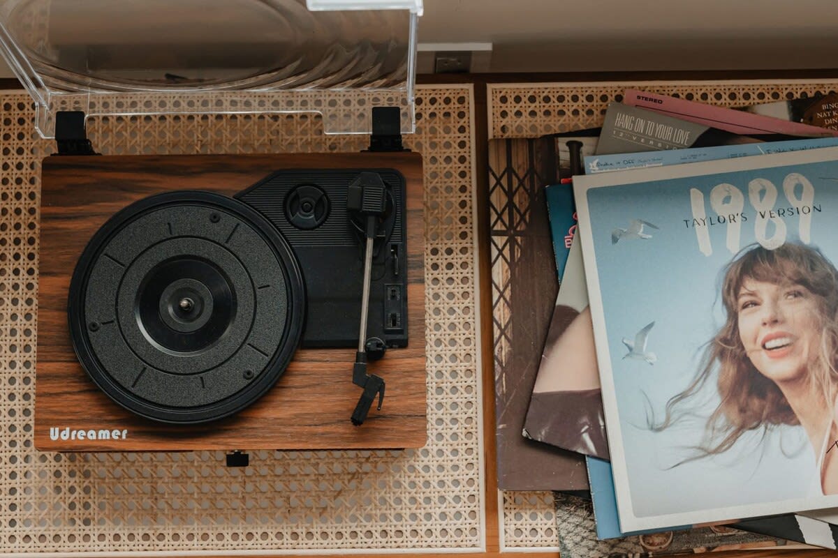 A close-up shot reveals a vintage-style wooden record player nestled within a light wood and cane media console, surrounded by a collection of vinyl records including Taylor Swift's "1989 (Taylor's Version)".