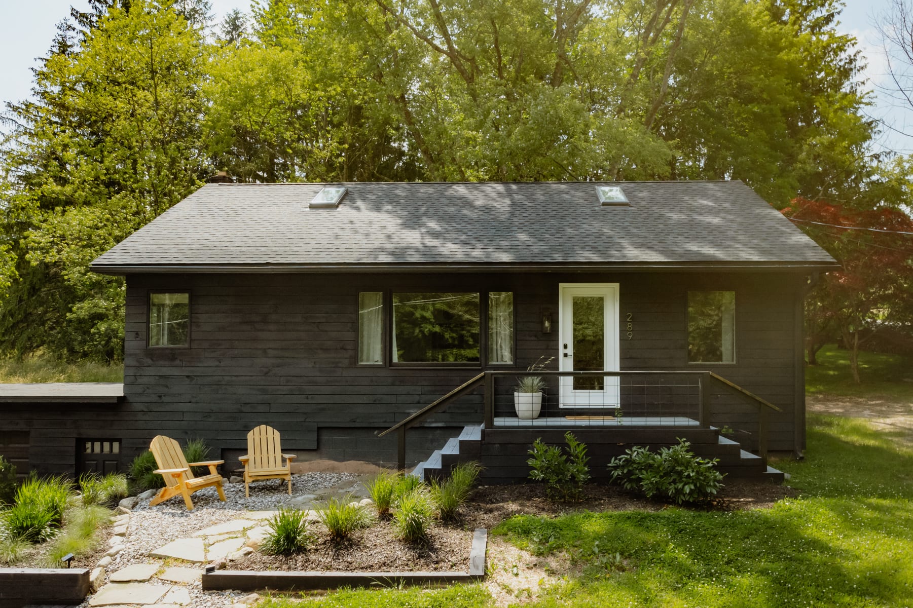 A modern black horizontal-sided house with a dark shingled roof and a white front door, flanked by large windows, presents a welcoming facade. A dark grey staircase with a white railing leads to the entrance, surrounded by green landscaping and two wooden Adirondack chairs.