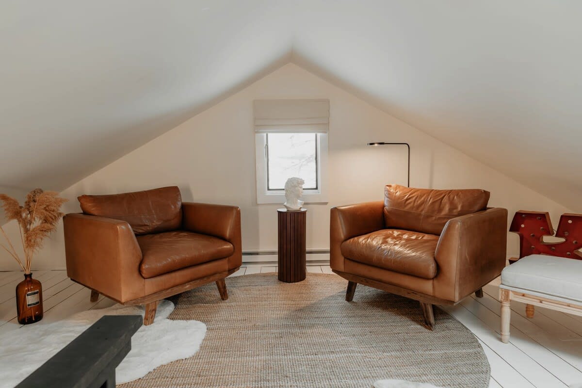 A bright living area with a sloped white ceiling showcases two comfortable brown leather armchairs on a round jute rug. A small window with a white blind is centered between the chairs, with a dark wood pedestal table holding a white decorative bust.