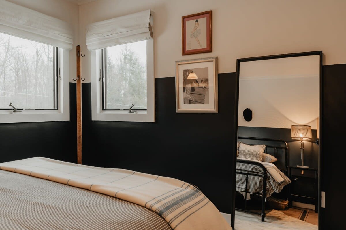 View of the two-tone bedroom with a black metal bed, striped bedding, and a full-length mirror reflecting the room. Two windows and a wooden coat rack are visible, enhancing the rustic modern aesthetic.