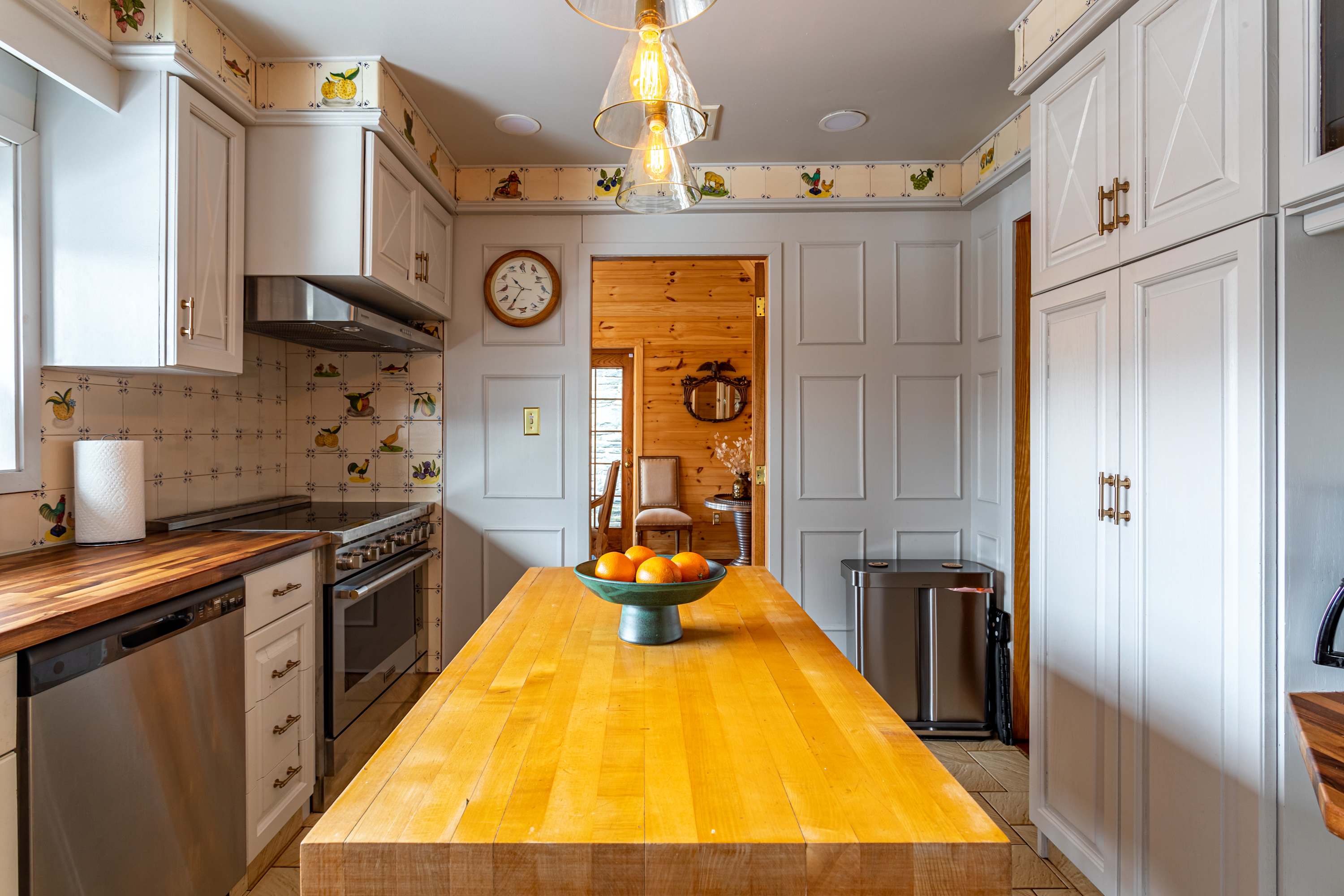 Another view of the kitchen, showcasing a stainless steel range with a hood, wooden countertops, and a large butcher block island with a bowl of oranges. The walls feature decorative wallpaper and white wainscoting.