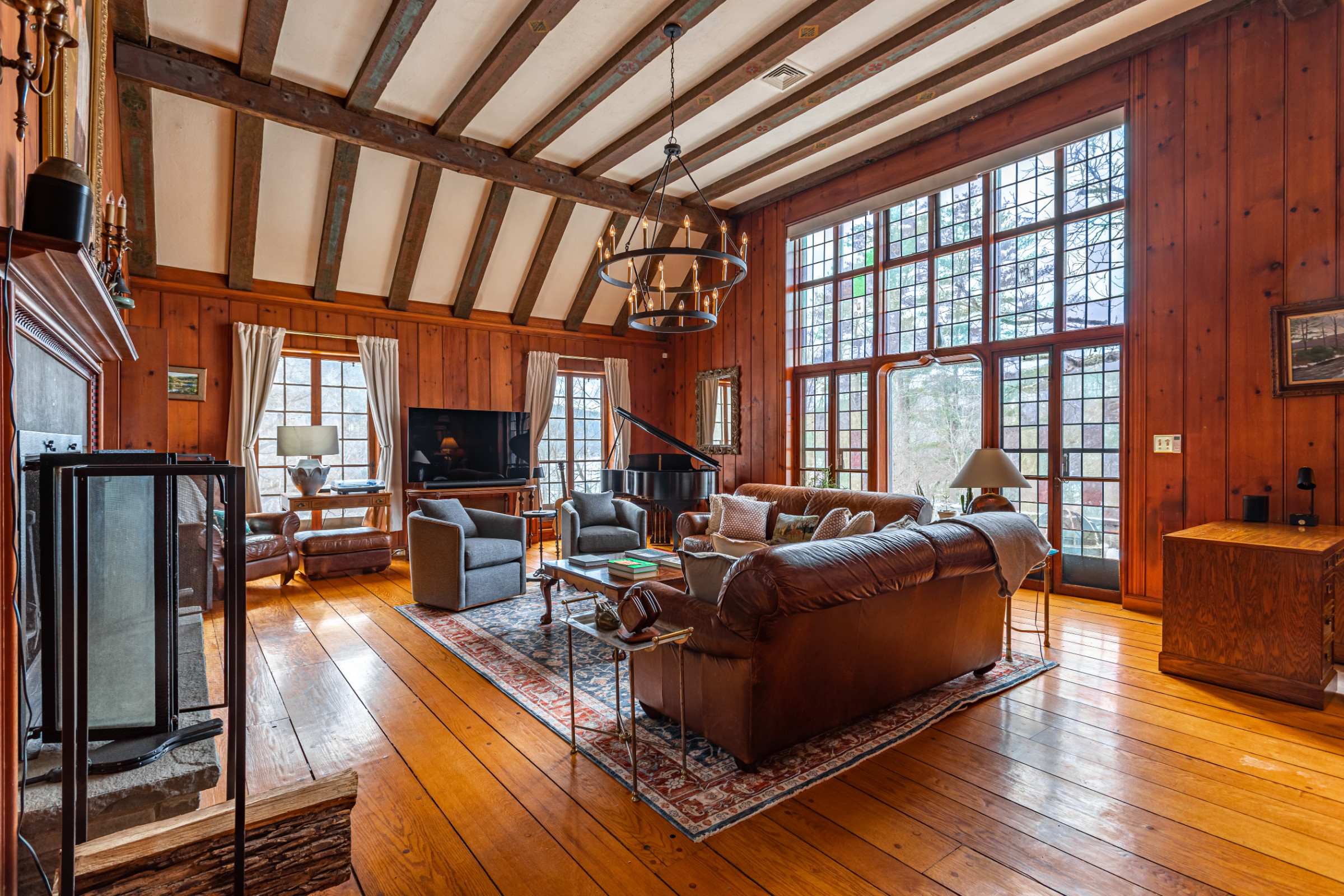 A spacious living room with high ceilings, exposed wood beams, and large windows, including a tall stained-glass window. Two brown leather sofas and two gray armchairs are arranged around a coffee table on a patterned rug.