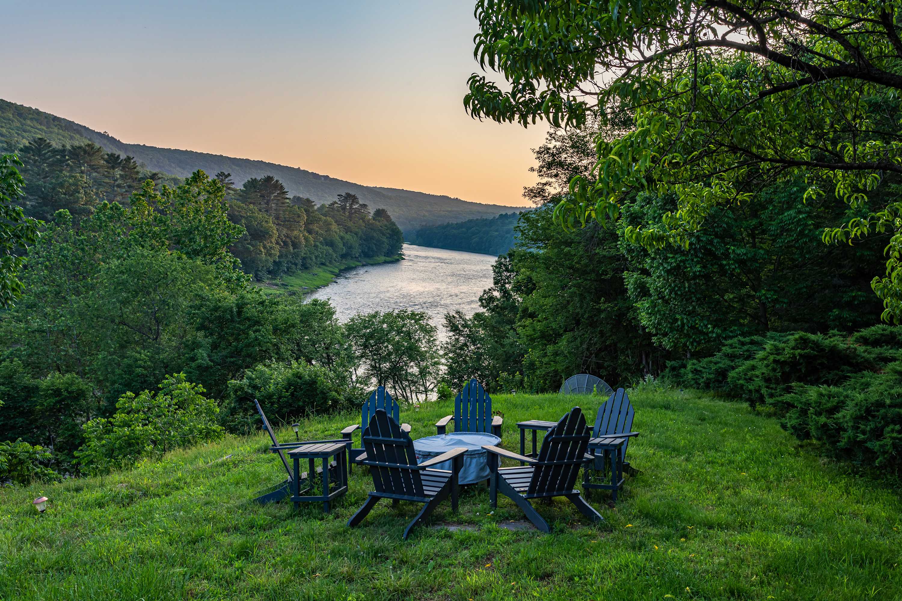 A serene outdoor fire pit area features dark Adirondack chairs arranged in a circle on a grassy hill, offering panoramic views of a wide river winding through lush green mountains under a soft sunset sky.