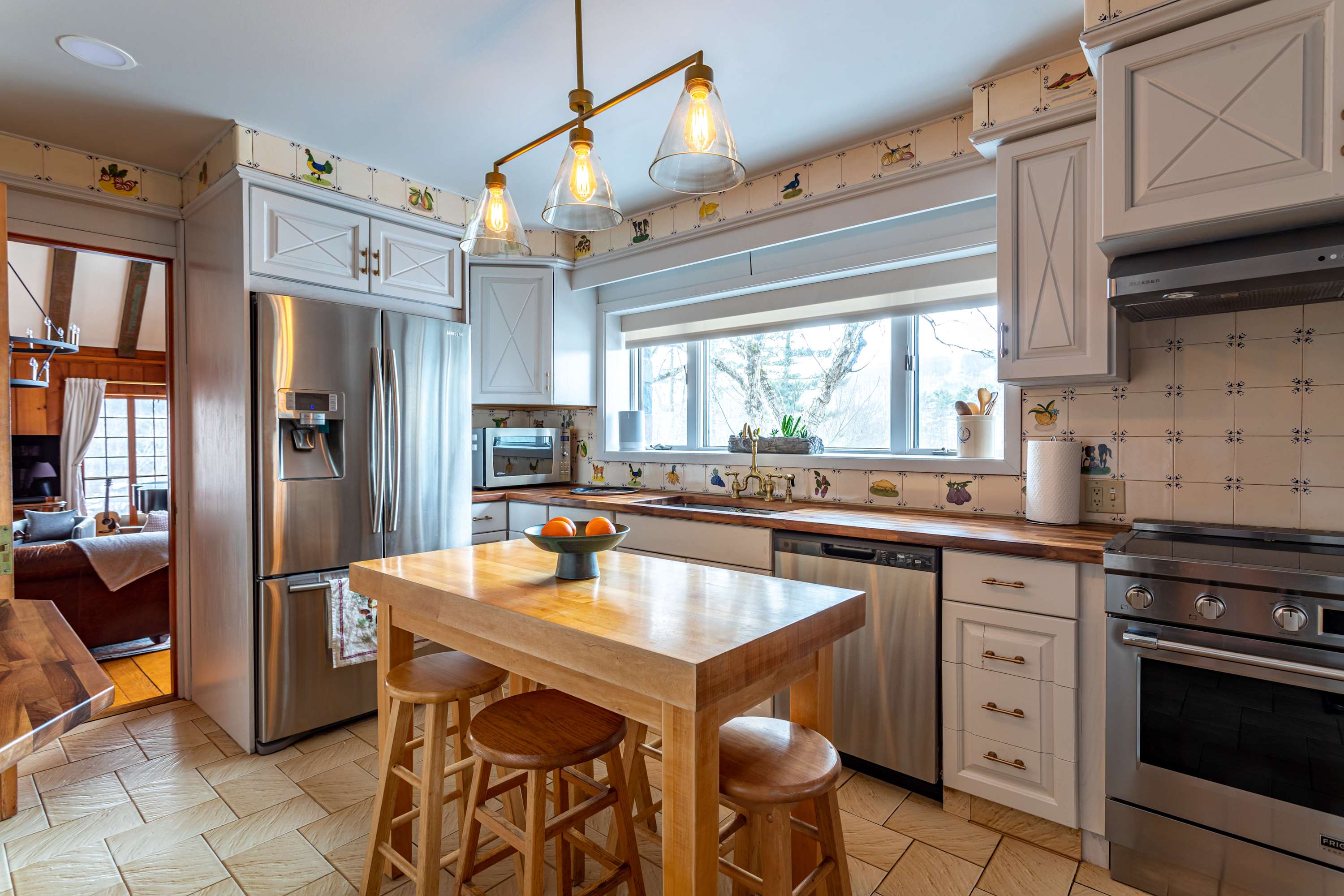 A bright kitchen with white shaker cabinets, butcher block countertops, and stainless steel appliances. A central wooden island with stools provides additional workspace and seating, illuminated by a modern pendant light.