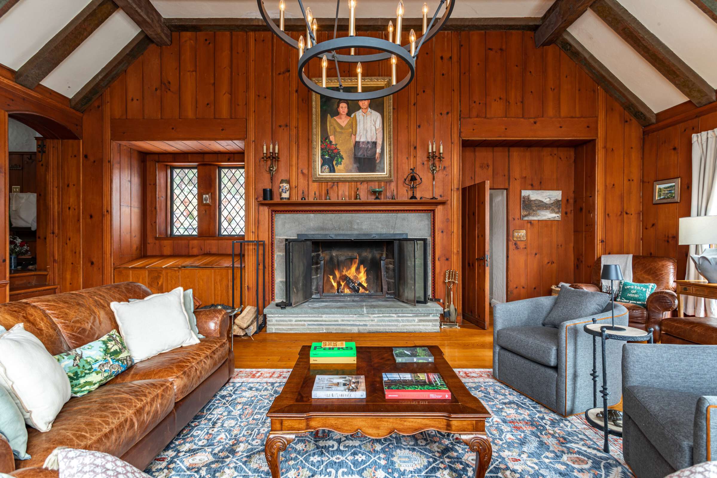 A warm living room features wood-paneled walls, a large stone fireplace with a roaring fire, and comfortable leather and upholstered sofas arranged around a wooden coffee table. A large chandelier hangs from the vaulted, beamed ceiling.