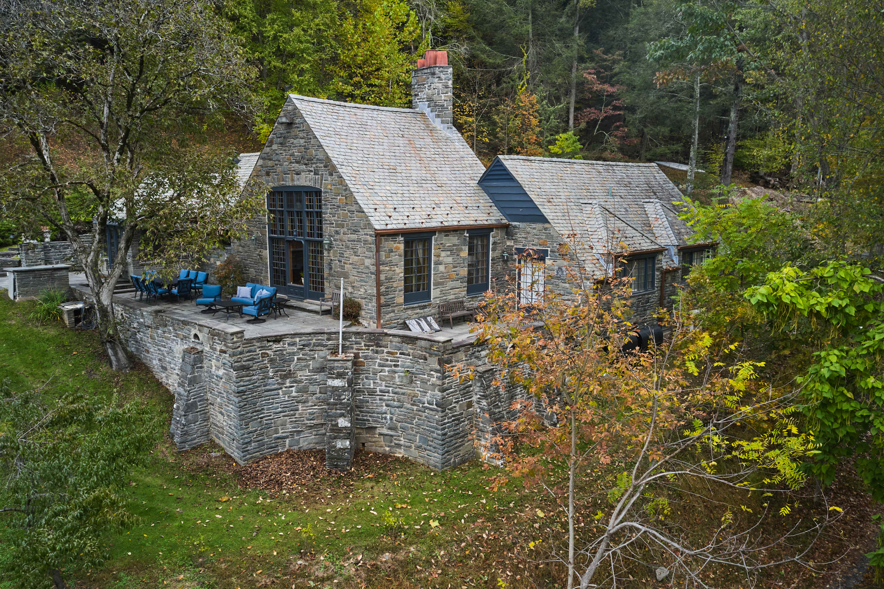 An aerial view showcases the rustic stone house with a multi-pitched slate roof, nestled into a hillside surrounded by dense autumn foliage, featuring a large patio with blue outdoor seating overlooking the landscape.