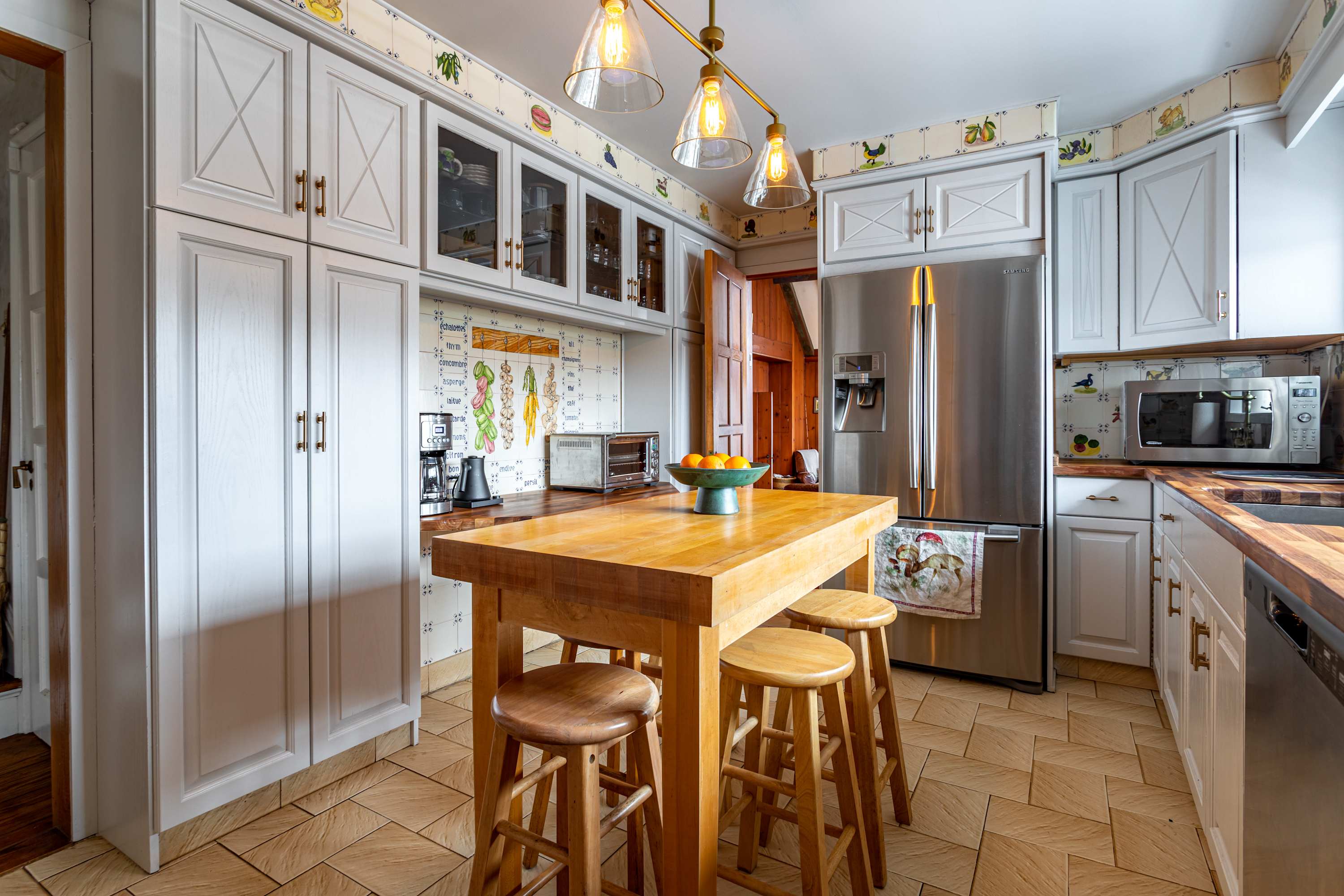 Bright kitchen featuring white shaker-style cabinets, a large wooden butcher block island with three stools, stainless steel appliances, and decorative wallpaper with fruit motifs. The floor is covered in light-colored tiles.