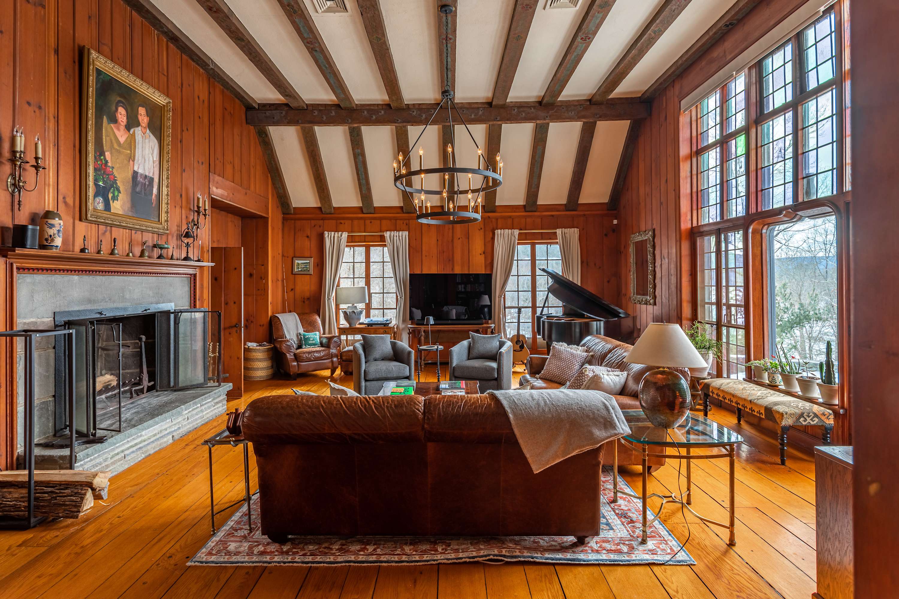 A wide view of the living room showcasing its high beamed ceiling, wood-paneled walls, and a large stone fireplace. Two brown leather sofas and two gray armchairs are arranged on a patterned rug, with a grand piano visible near the large windows.
