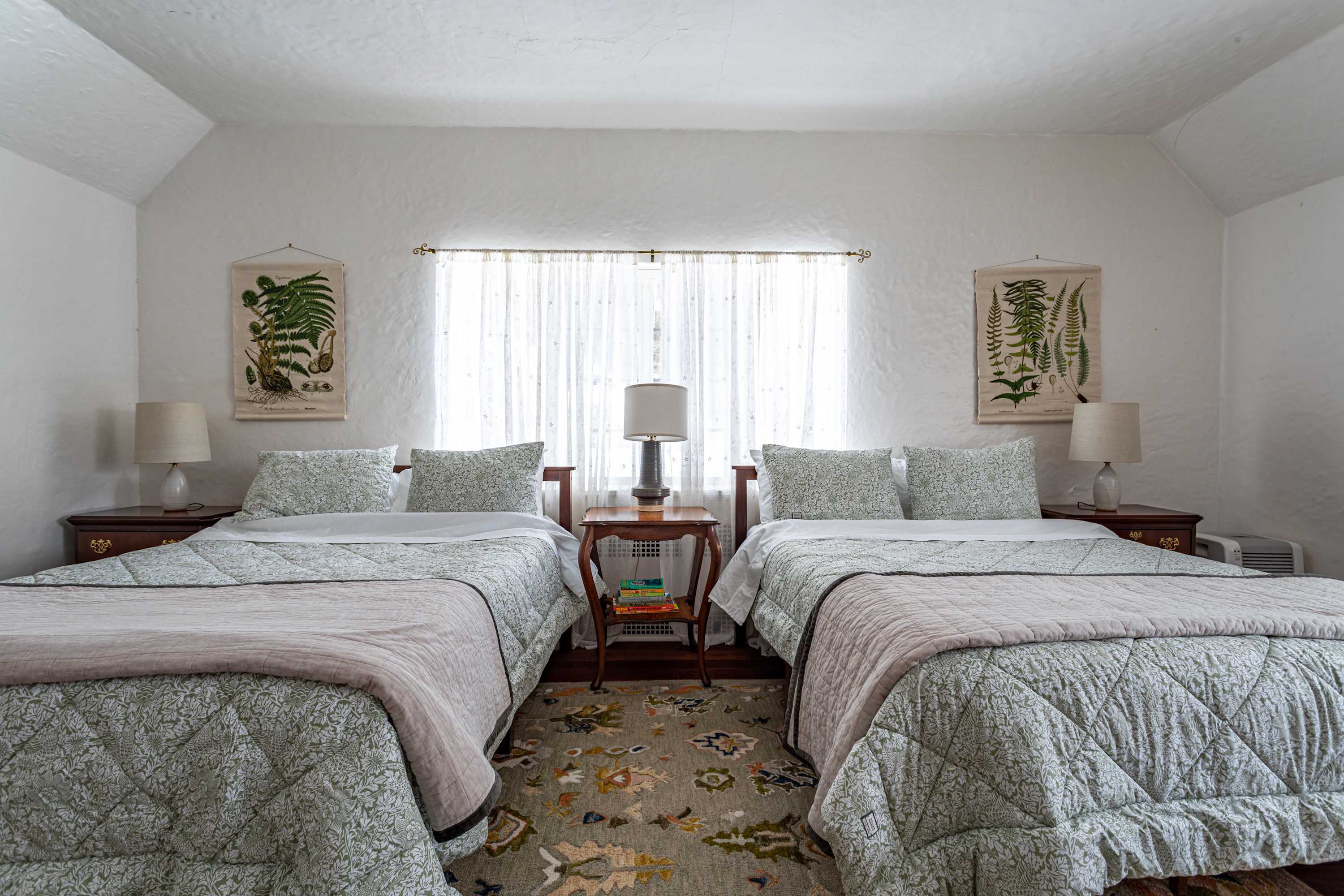 A bright bedroom featuring two twin beds with light green patterned quilts, a shared wooden nightstand with a lamp, and large windows covered by sheer white curtains.
