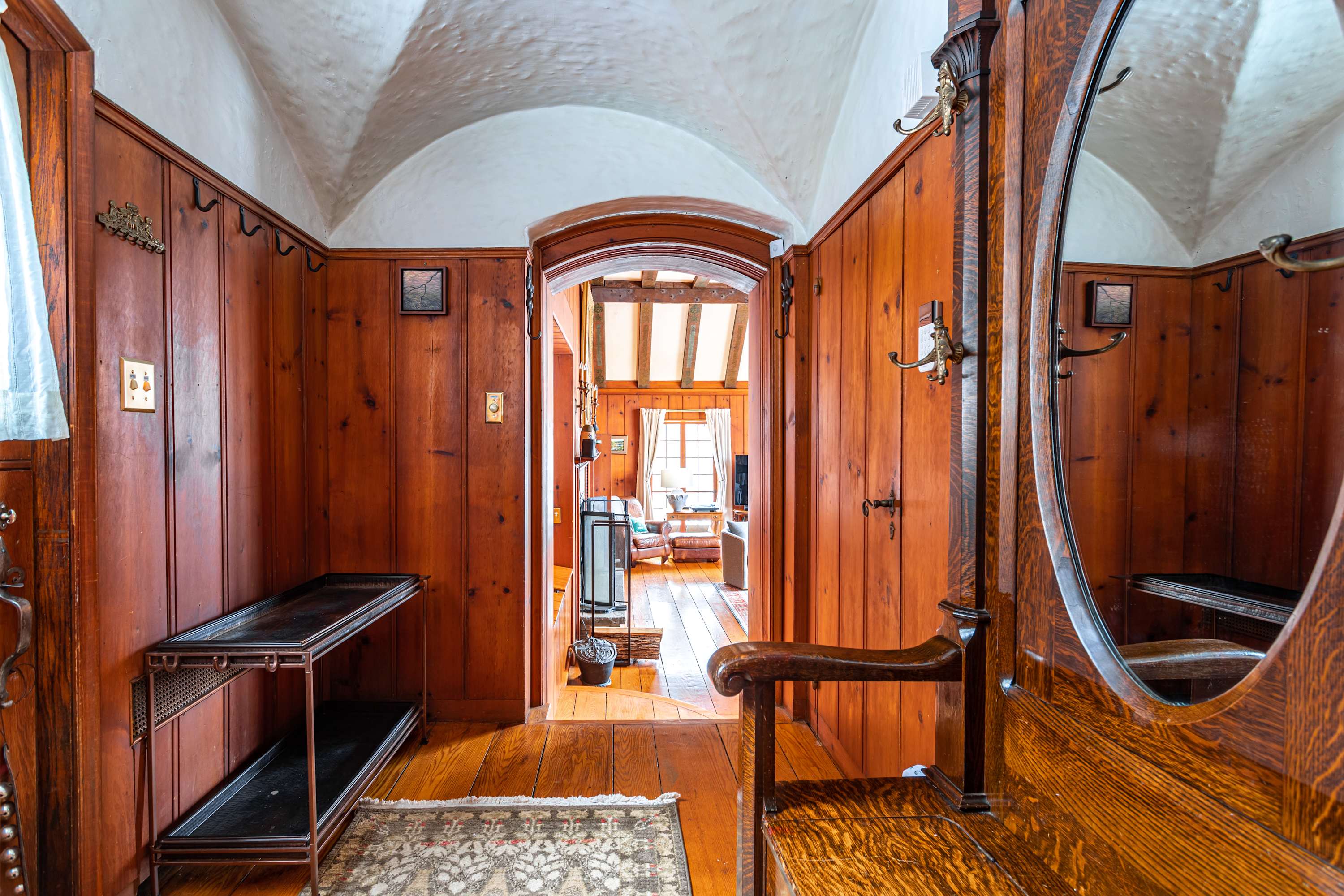 A warm, inviting foyer with rich wood paneling on the walls and a vaulted white ceiling. A large wooden hall tree with a mirror stands to the right, and a dark console table is on the left, leading to an arched doorway into another room.