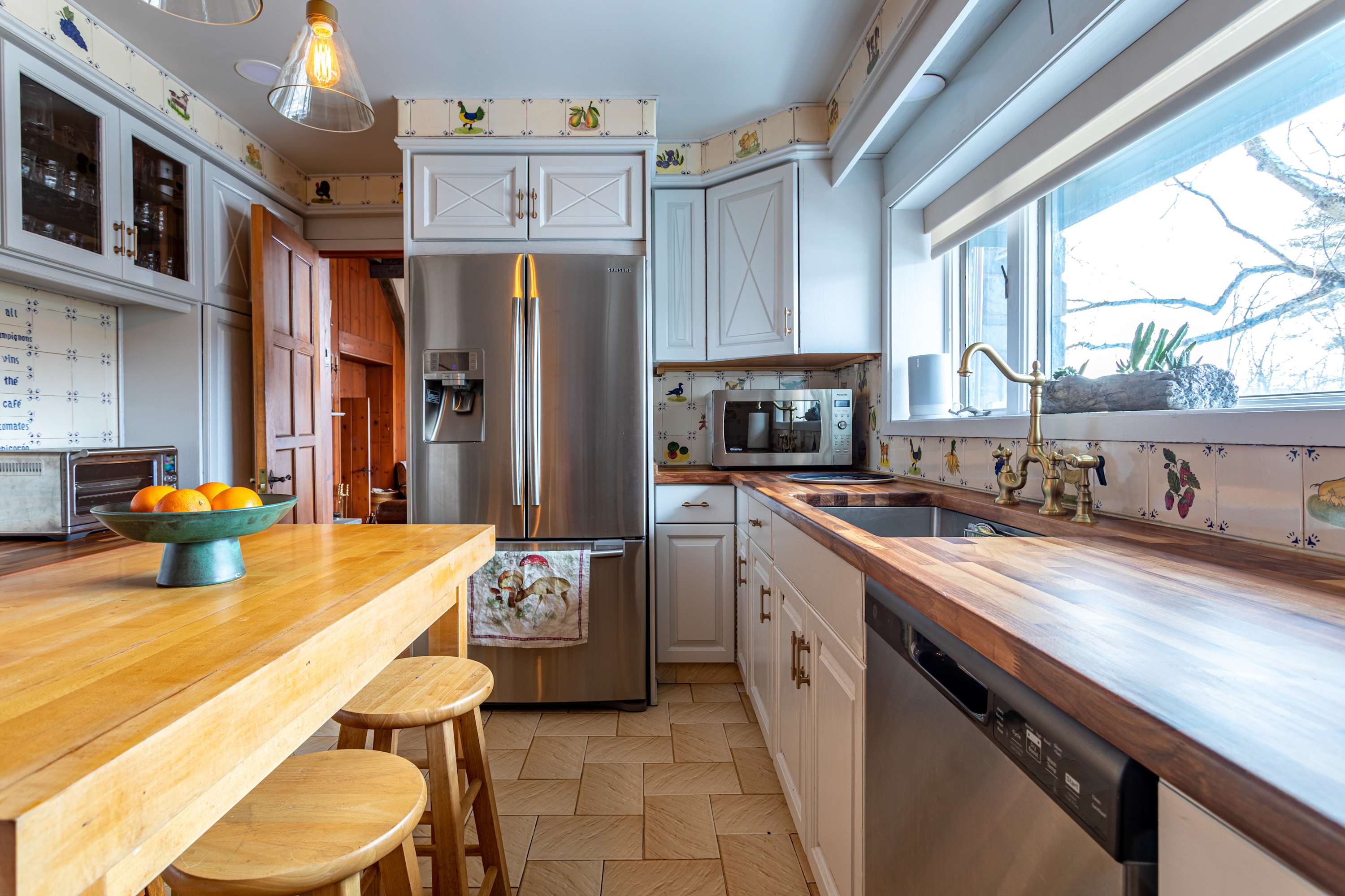 A close-up view of the kitchen showing the stainless steel dishwasher, sink with a brass faucet, and butcher block countertops. White shaker cabinets line the walls, and a wooden island with stools is visible in the foreground.