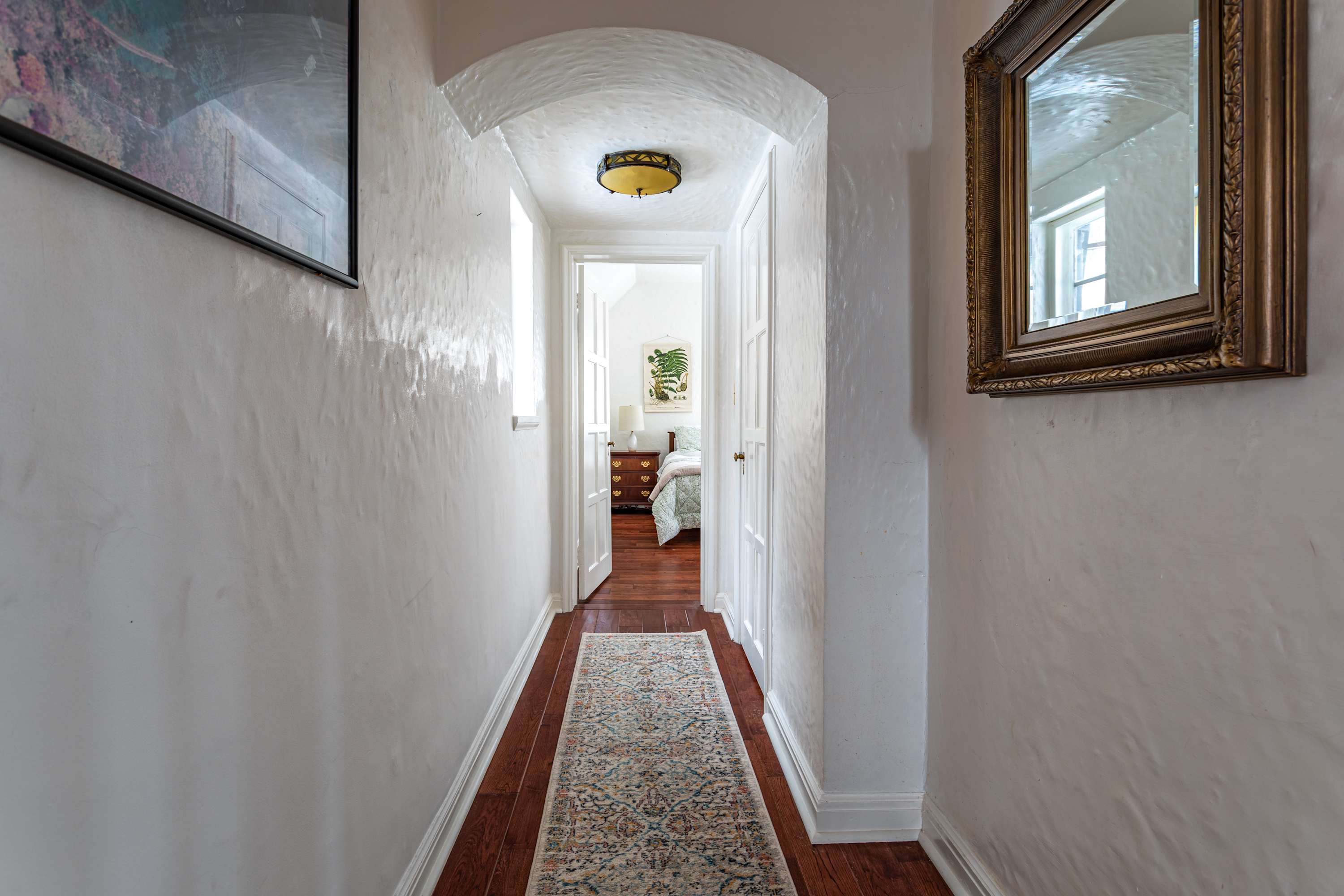 A narrow hallway with white textured walls, dark hardwood floors, and a patterned runner rug, leading to an arched doorway revealing a glimpse of a bedroom.