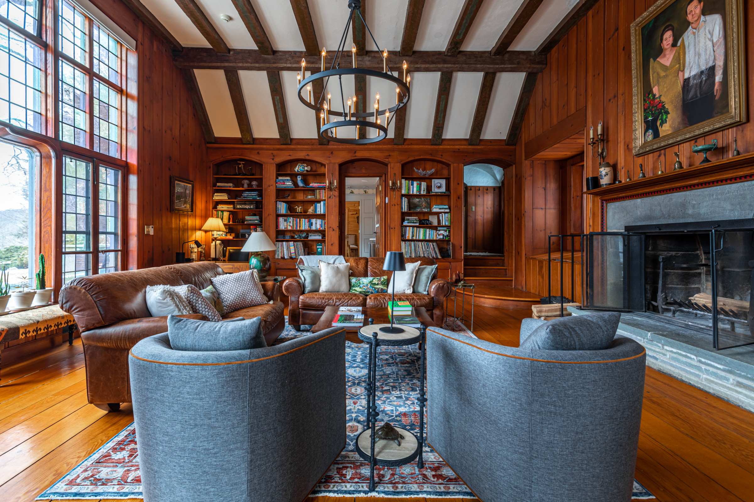 A comfortable living room with rich wood paneling, exposed ceiling beams, and built-in bookshelves filled with books and decor. Two brown leather sofas and two gray armchairs are arranged on a patterned rug, facing a stone fireplace.