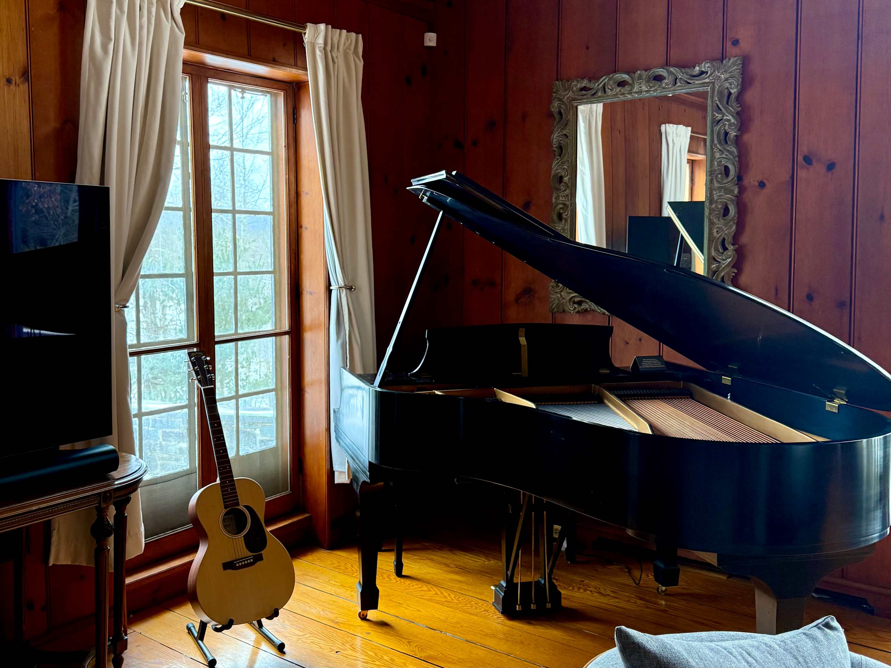 A black grand piano and an acoustic guitar stand by a tall window with white curtains, showcasing the musical amenities within the wood-paneled living room. A decorative mirror reflects the room's elegant details.