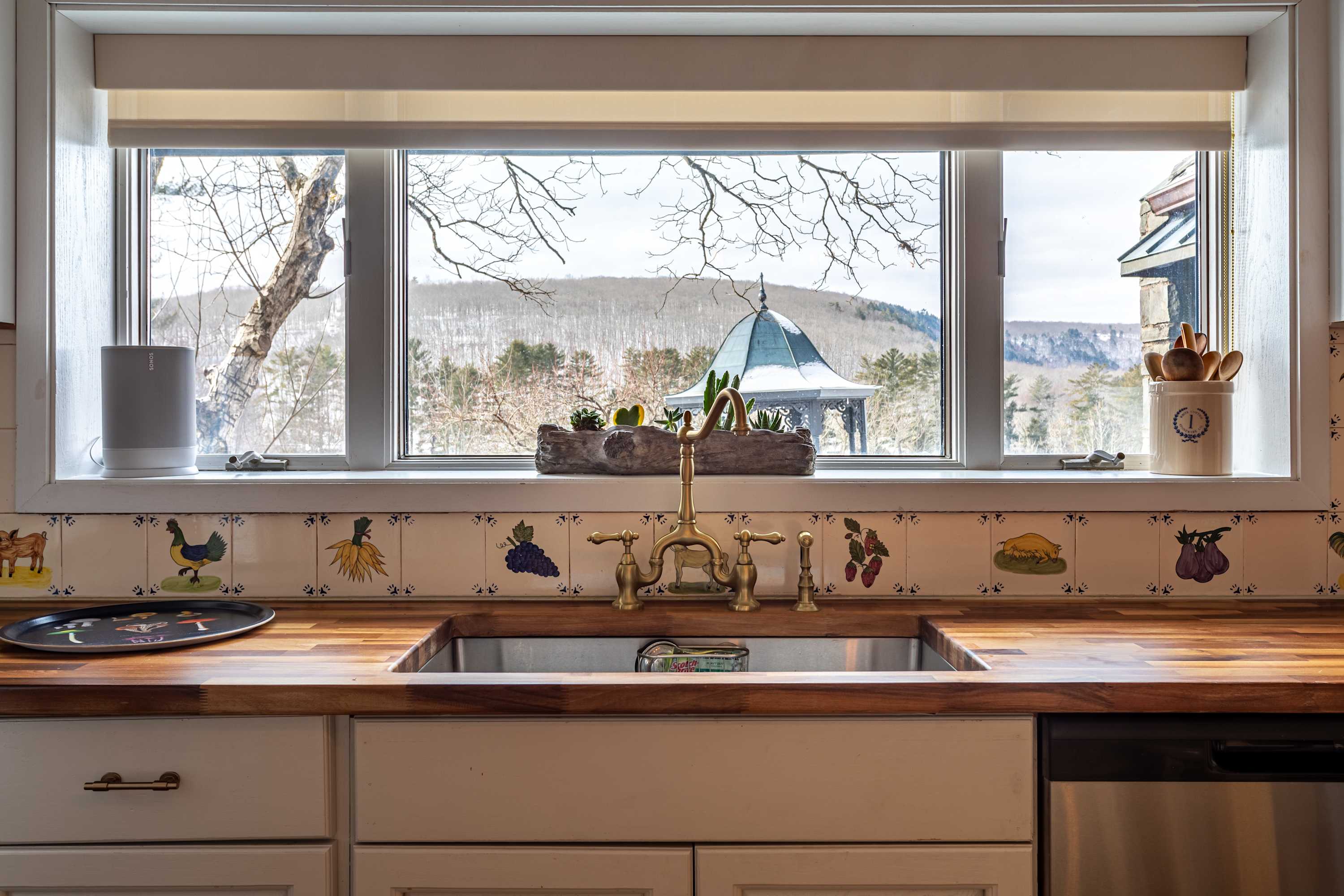 A kitchen sink with elegant brass fixtures is set into a wooden countertop, complemented by a decorative tiled backsplash. A large window offers a scenic view of a snowy, tree-covered landscape and a distant gazebo.
