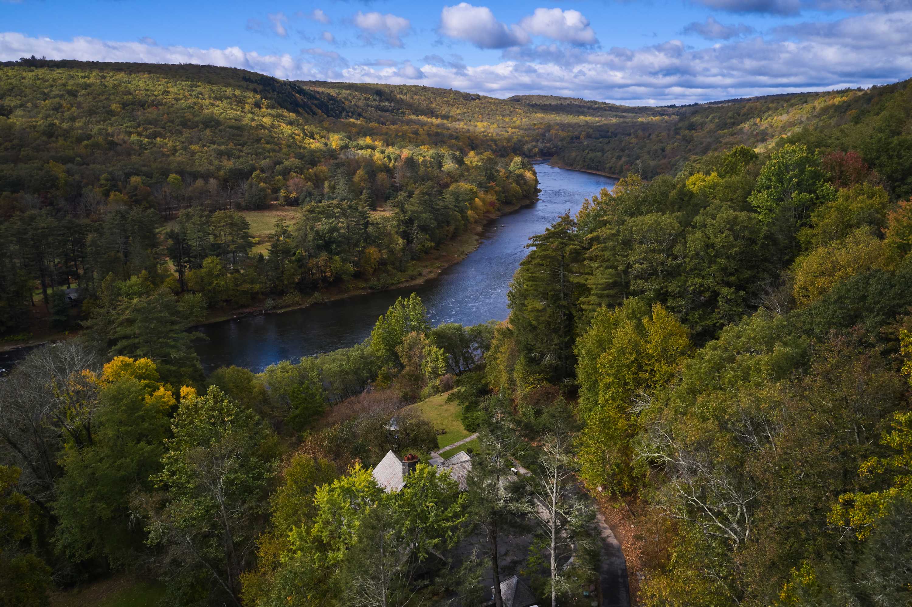 An aerial perspective revealing a house situated near a winding river, surrounded by dense, colorful autumn forests and distant mountains under a bright blue sky.