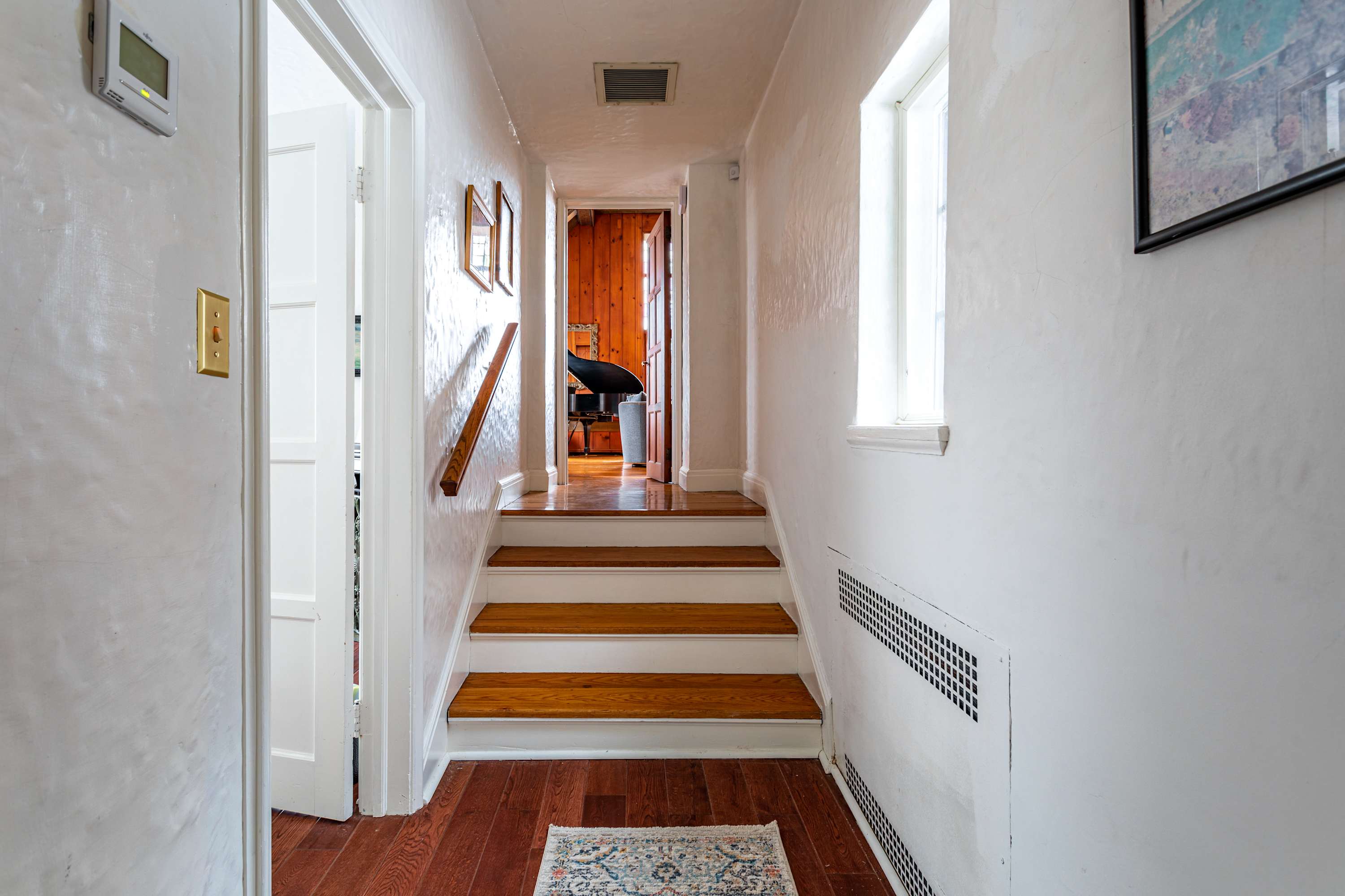 Narrow hallway with textured white walls and a wooden staircase leading up, featuring a wooden handrail. A small window provides natural light, and a patterned rug lies on the hardwood floor.