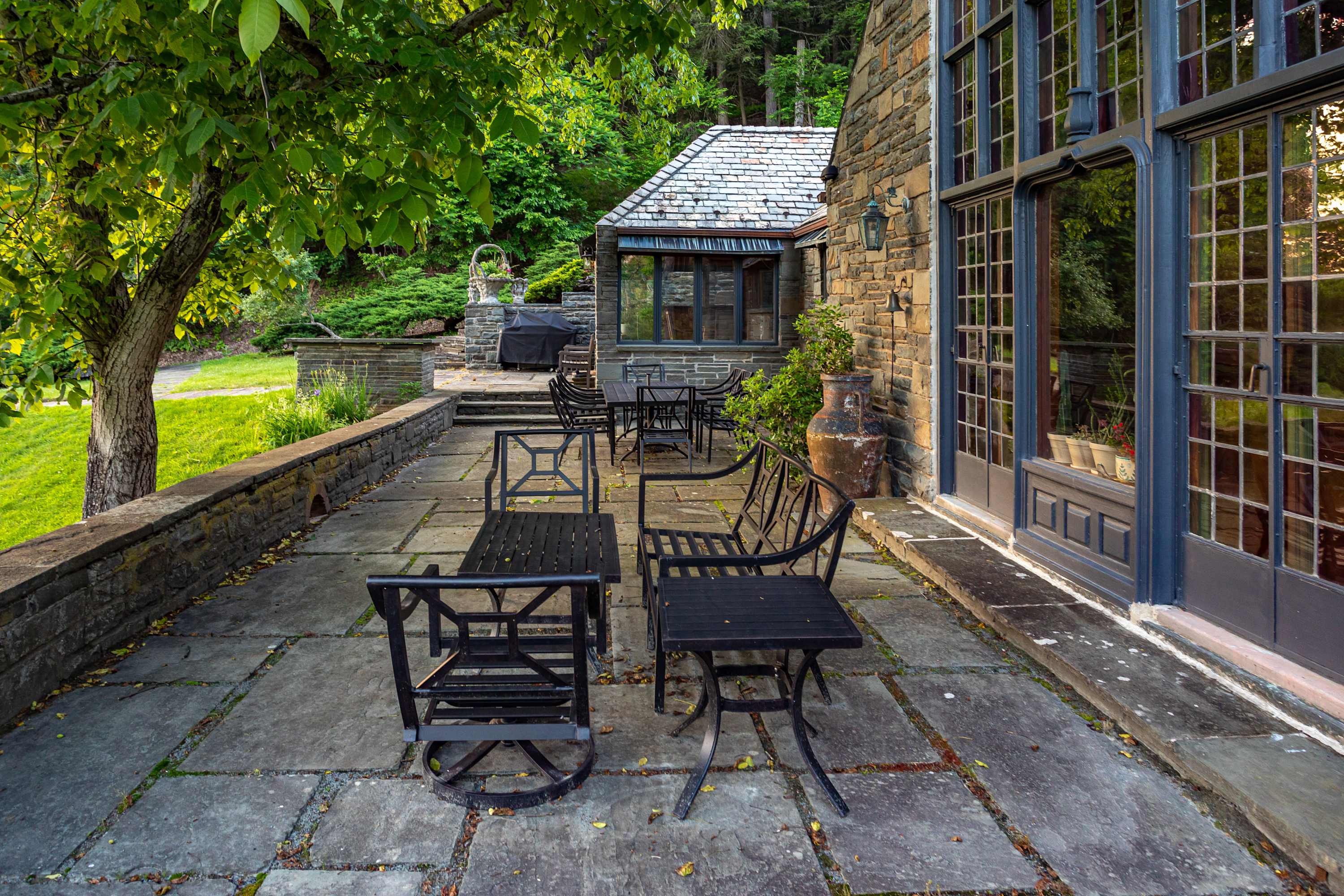 An expansive stone patio features multiple black metal outdoor dining sets, including a smaller bistro set and a larger table with chairs, set against a rustic stone house with large windows and surrounded by lush greenery.