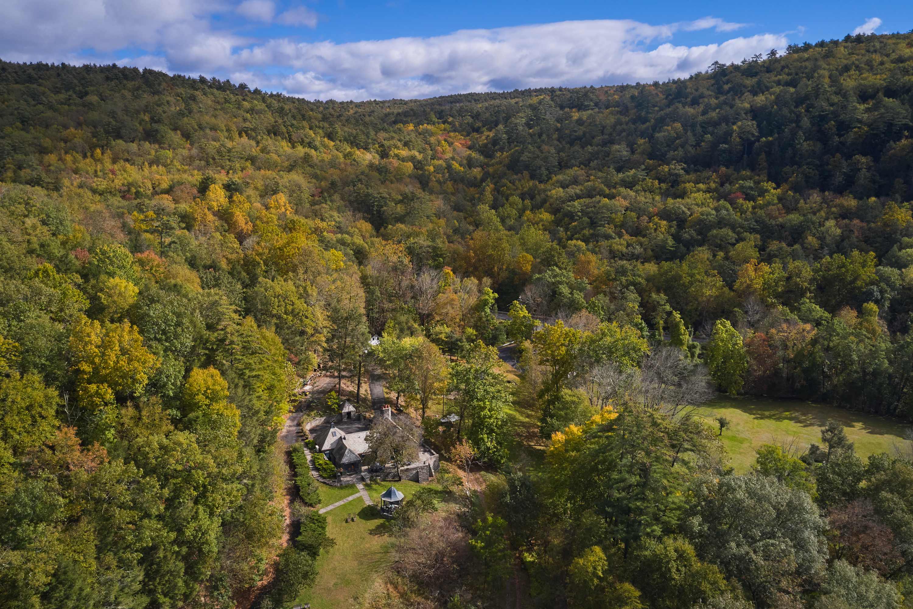 An aerial view showcasing a stone house nestled among vibrant green and yellow autumn trees, surrounded by rolling hills and mountains under a blue sky with scattered clouds.