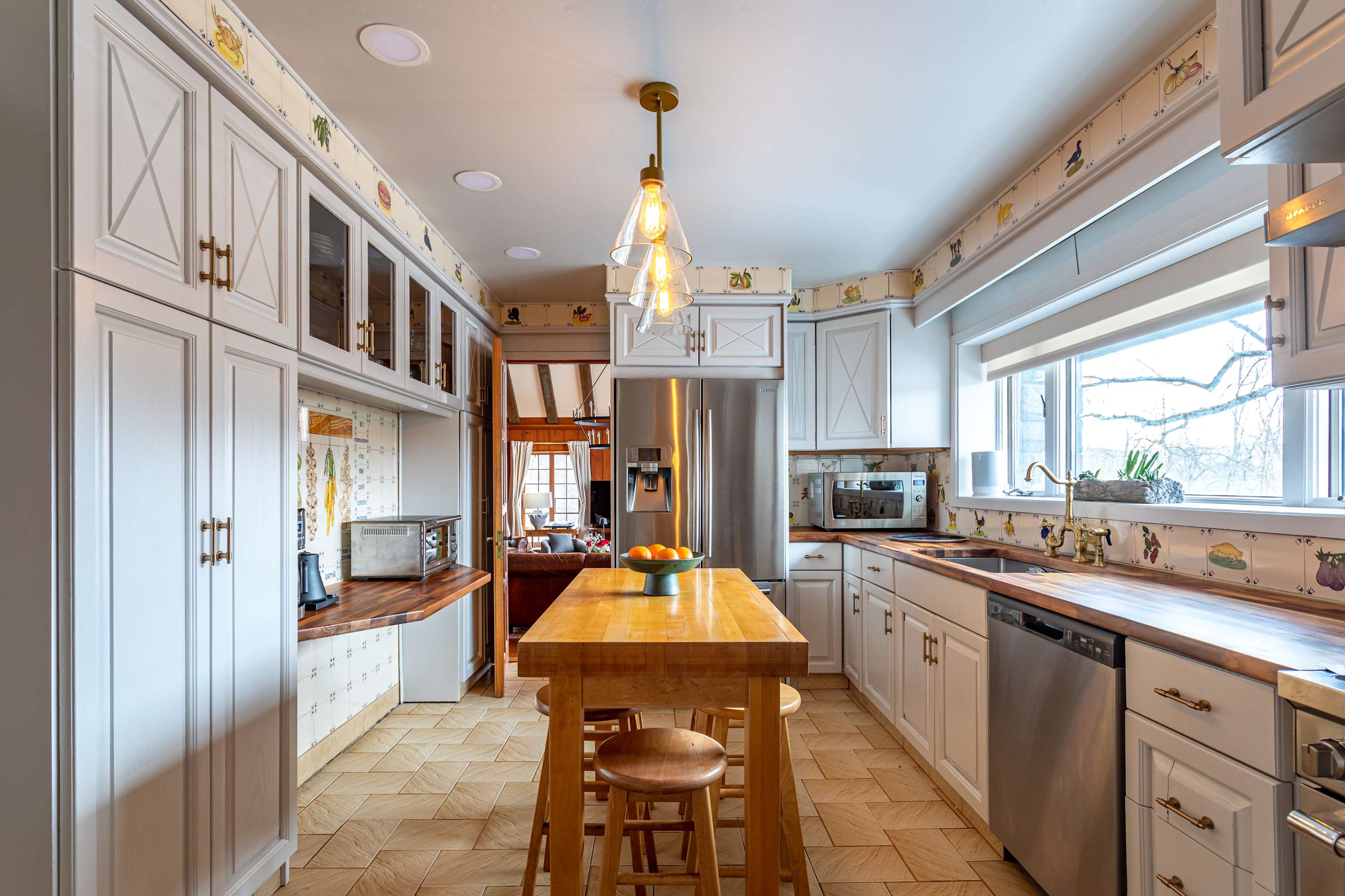 A galley-style kitchen featuring white shaker cabinets, butcher block countertops, and stainless steel appliances, including a large refrigerator. A wooden island with stools is centrally placed, and a large window overlooks the outdoors.