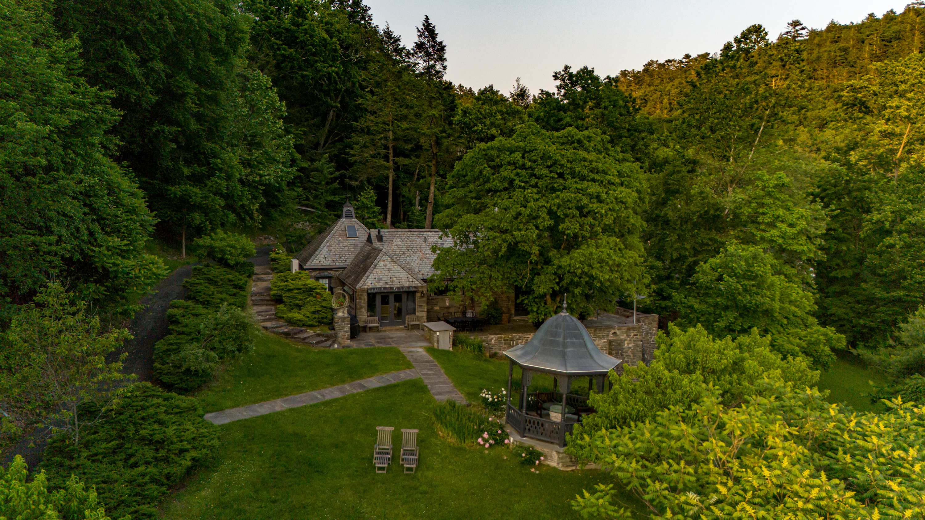 An aerial view showcases the charming stone house with its slate roof, a winding stone pathway, a dark gazebo, and a well-maintained grassy yard, all enveloped by dense, vibrant green trees and foliage.
