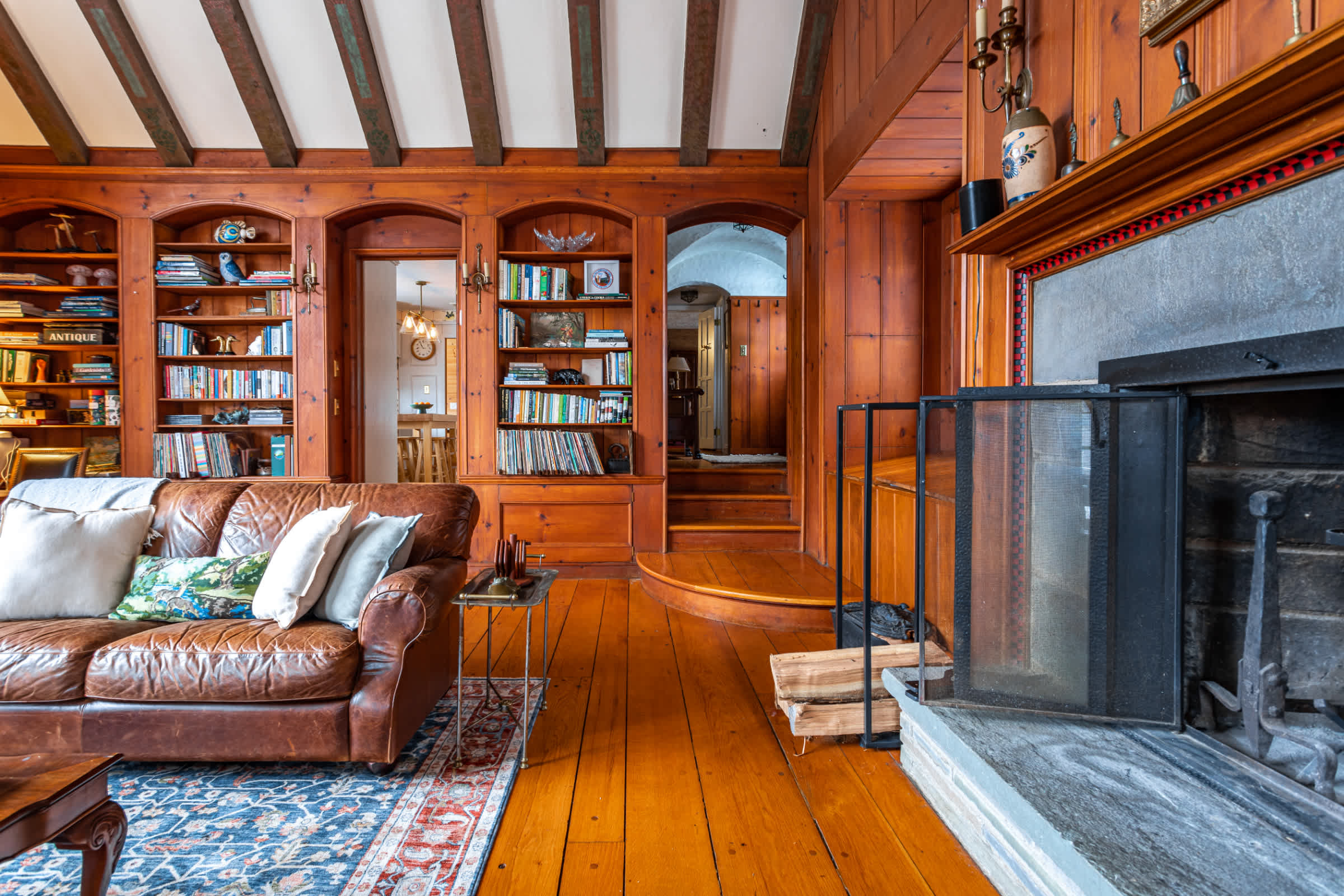 A cozy living room featuring extensive wood paneling, exposed ceiling beams, and a large stone fireplace with a metal screen. A brown leather sofa with throw pillows sits on a patterned rug, facing the fireplace.