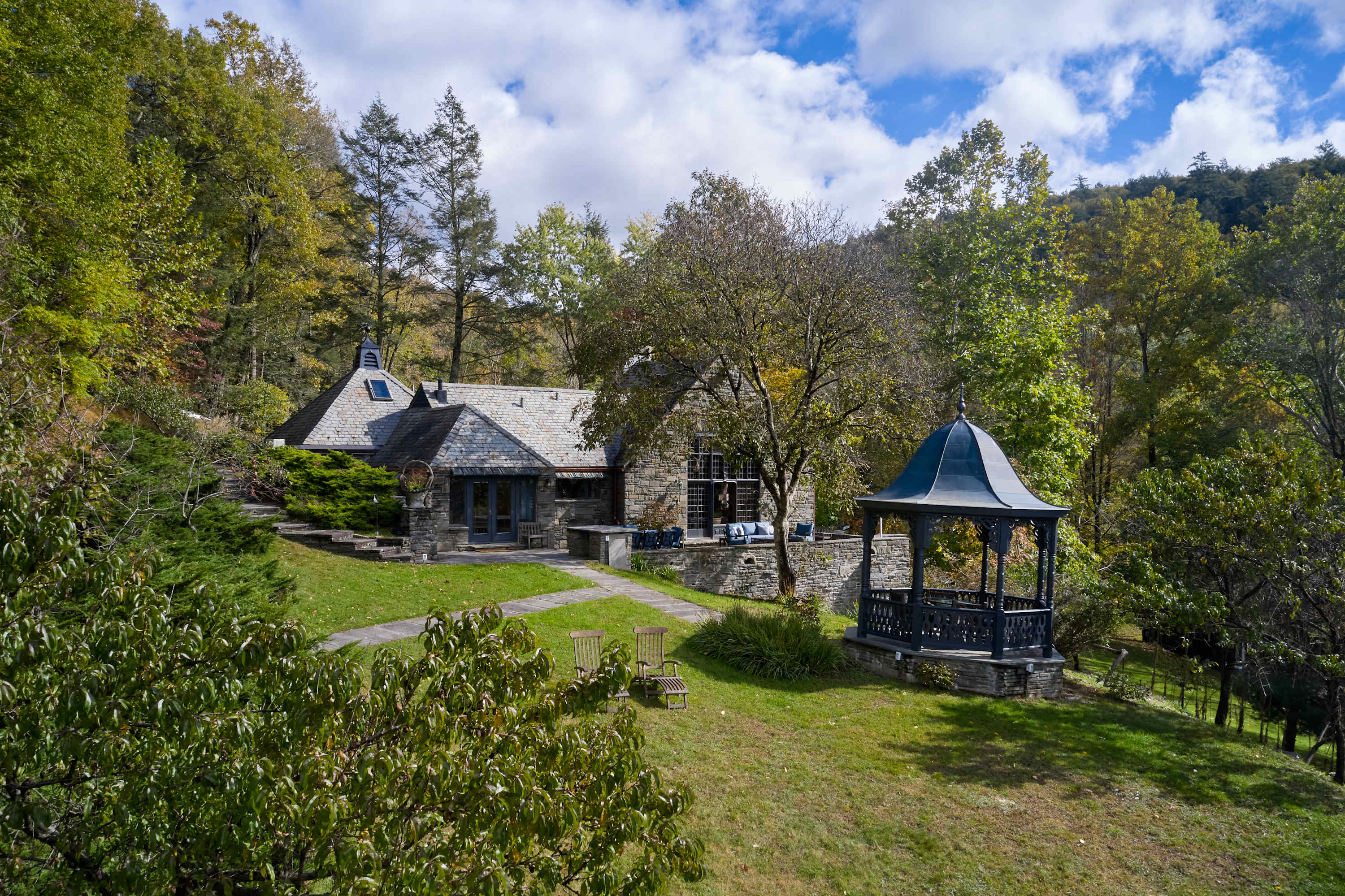 An expansive aerial view captures the stone house with its slate roof, a winding stone pathway, a dark gazebo, and a large grassy yard, all surrounded by dense green trees and foliage.