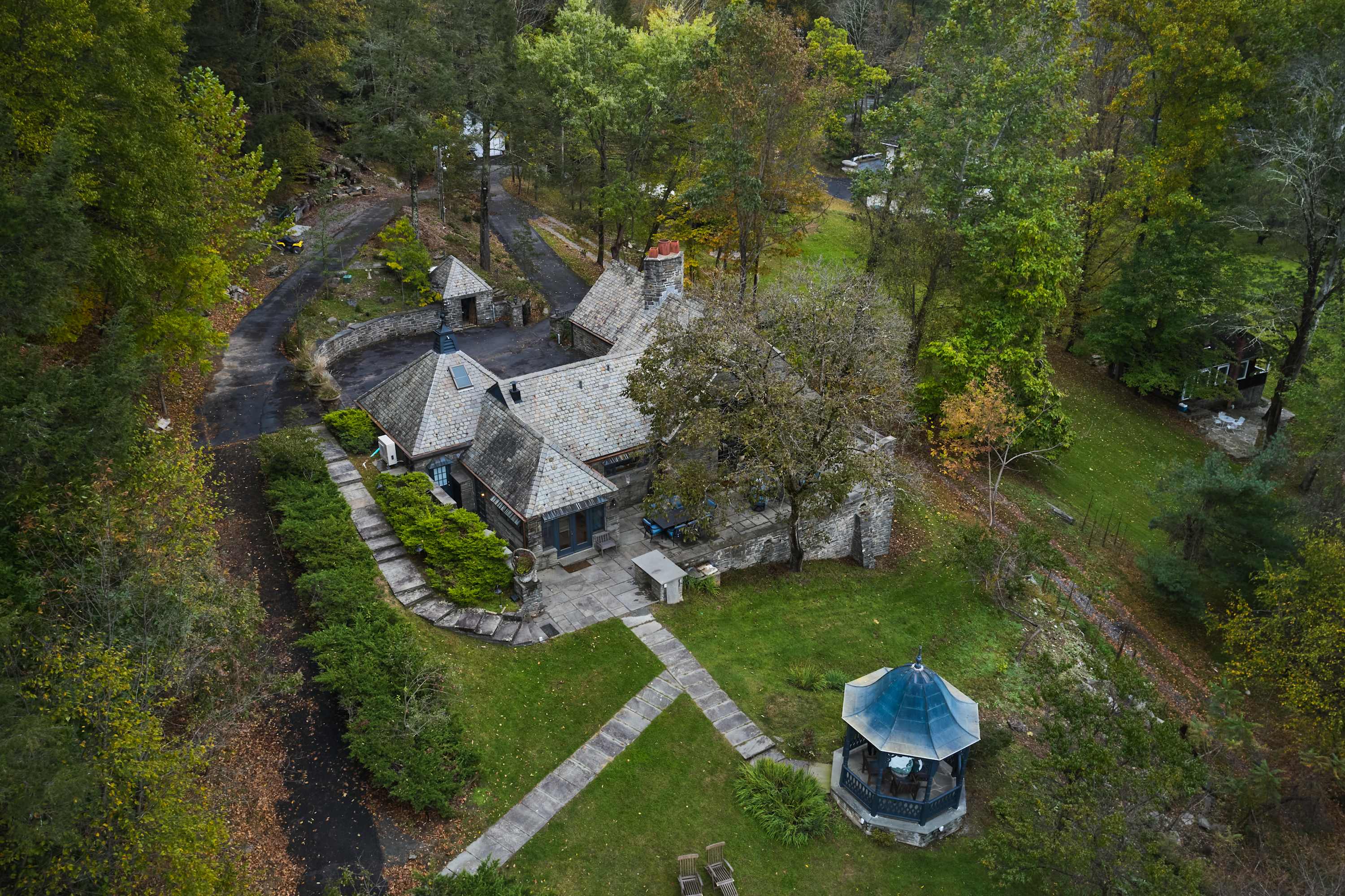 An overhead view of a unique stone house with a multi-gabled roof, surrounded by green lawns, mature trees, a paved driveway, and a small gazebo in the foreground.
