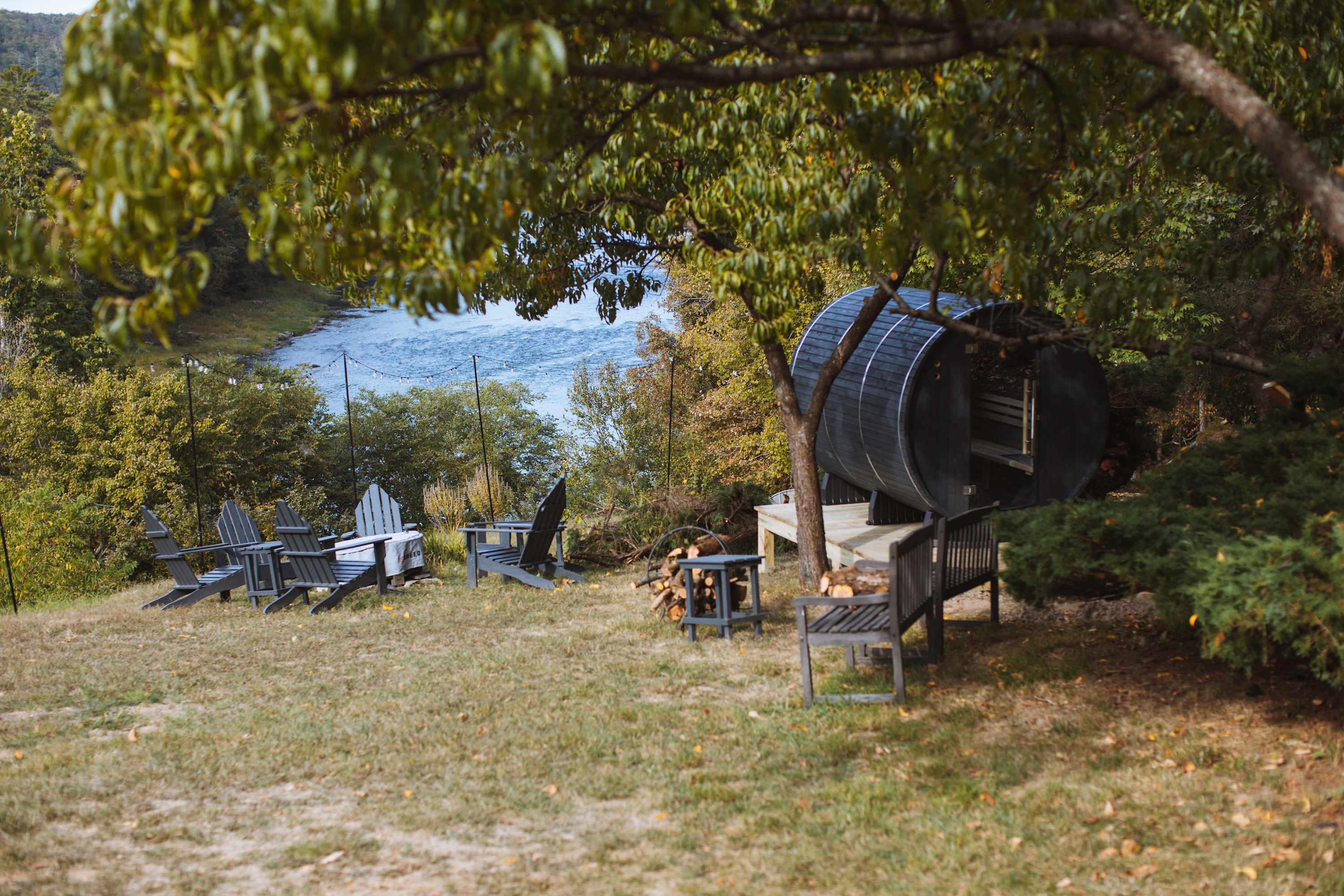 A barrel sauna sits on a wooden deck in a grassy yard, accompanied by several Adirondack chairs and a pile of firewood, all overlooking a scenic river through the trees, creating a perfect relaxation spot.