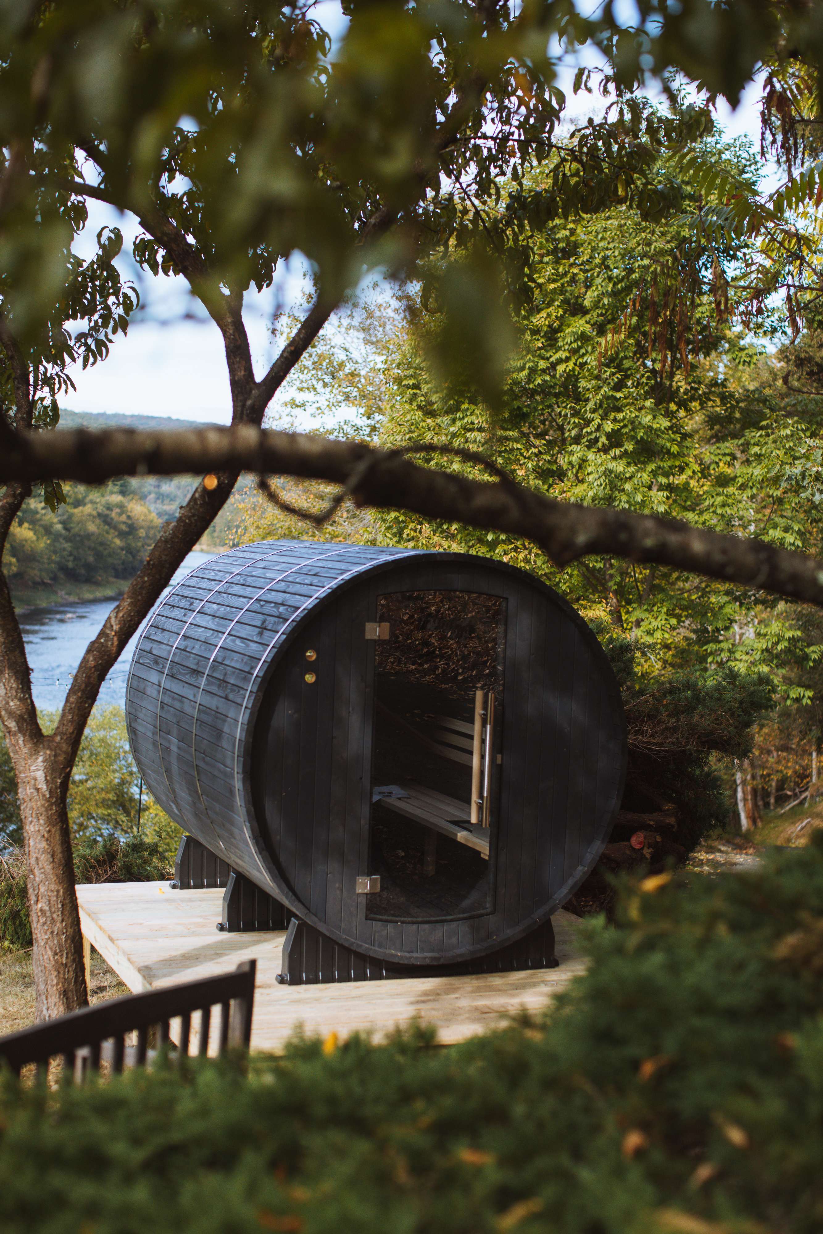A dark, barrel-shaped sauna with a glass door sits on a wooden deck overlooking a river, framed by tree branches and lush foliage, offering a serene outdoor wellness experience.