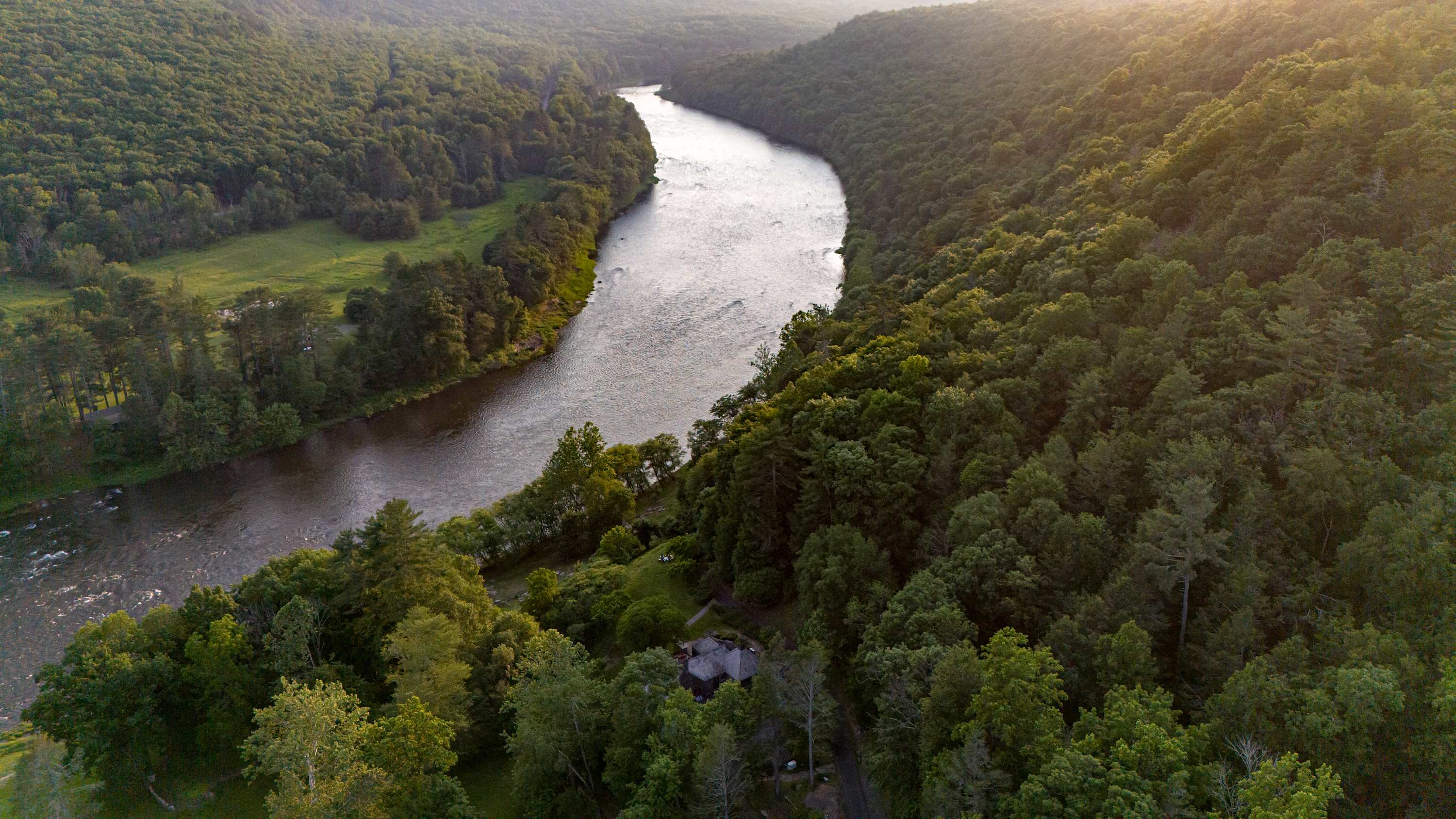 A high aerial view captures a wide river meandering through a vast, lush green forest, with a stone house subtly nestled among the trees on the riverbank, all under a soft, golden light.