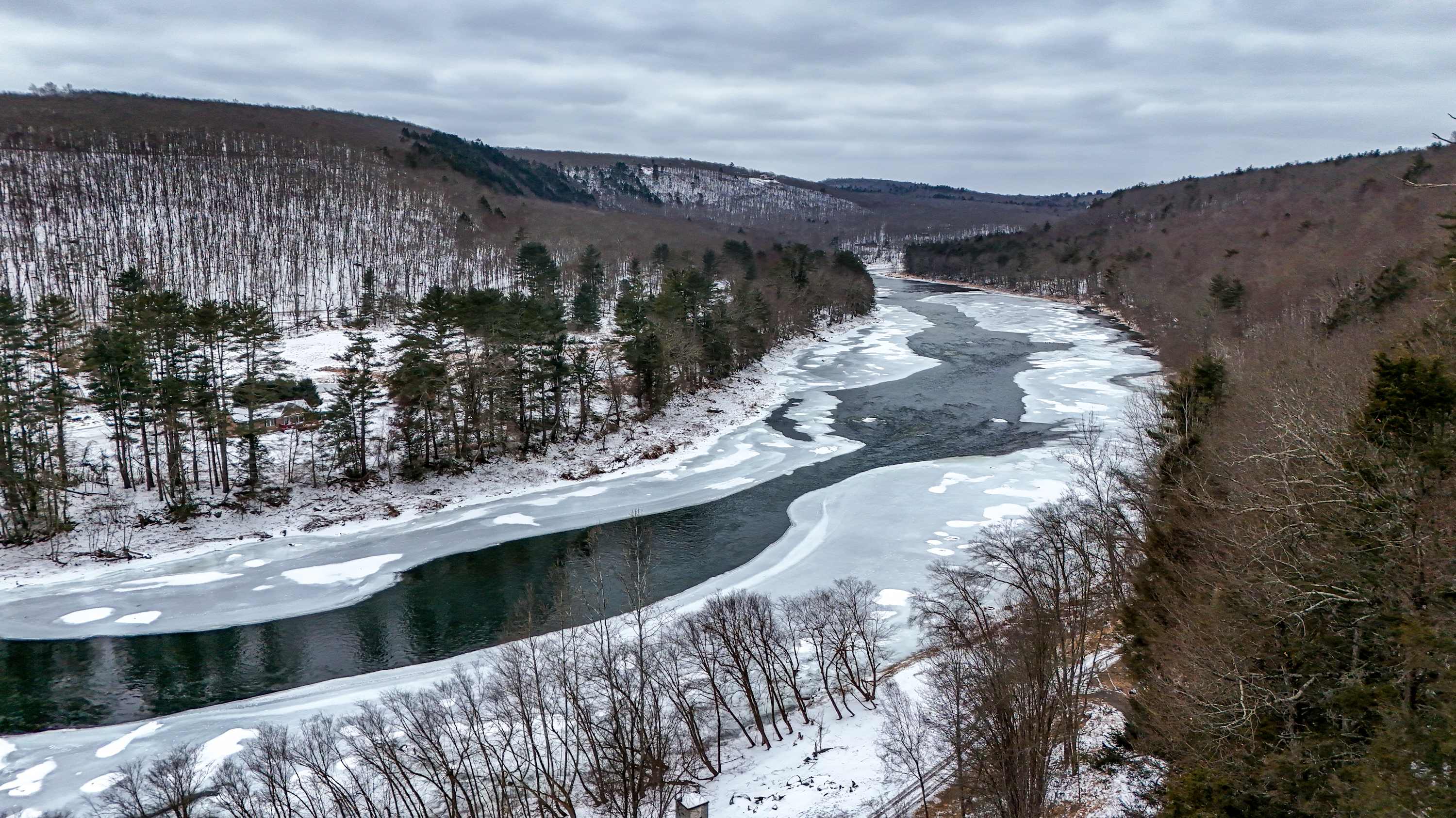 An aerial view reveals a wide river partially covered in ice and snow, winding through a valley flanked by snow-dusted hills and bare winter trees under a cloudy sky.