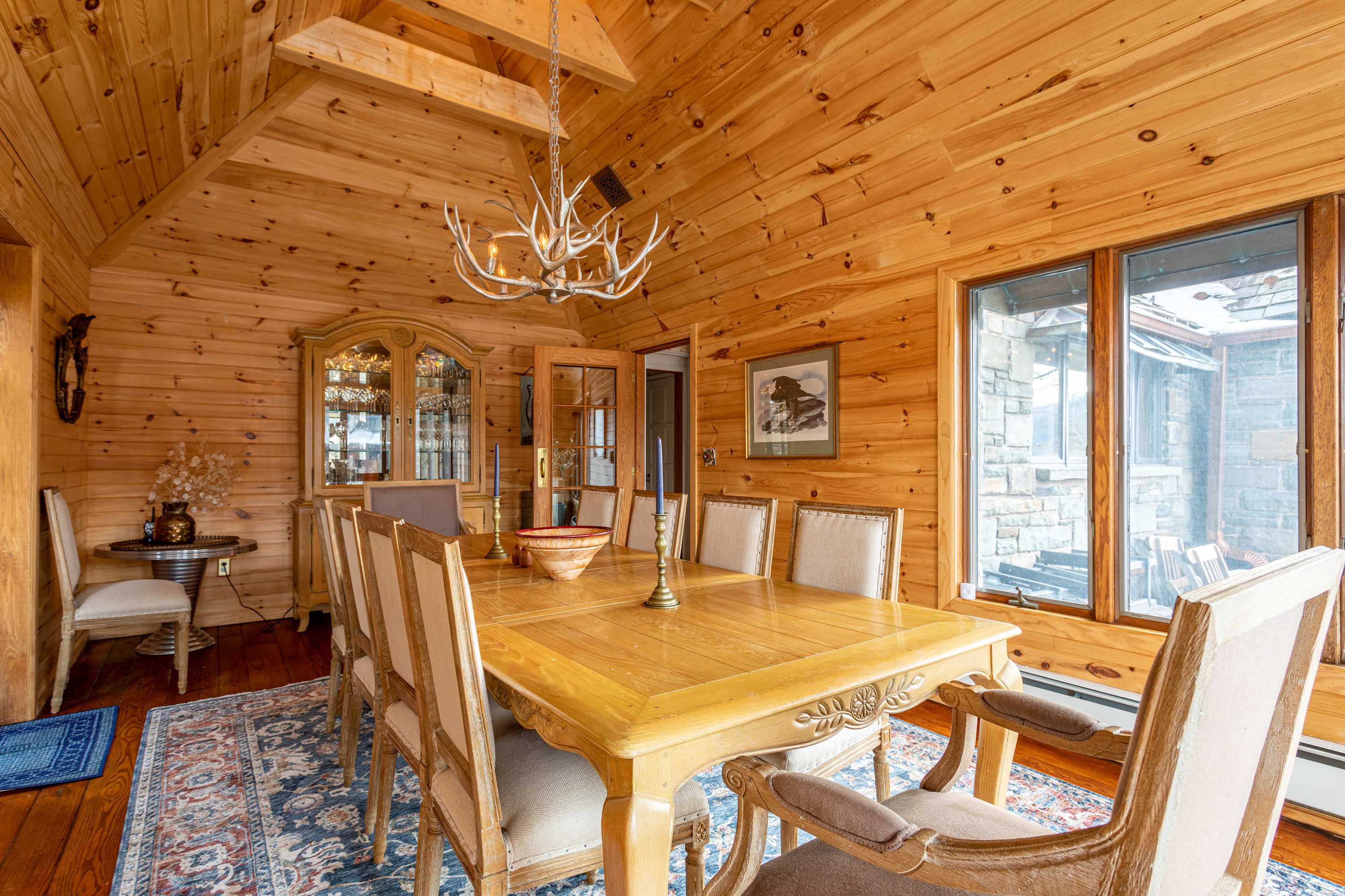 Rustic dining room with light wood paneled walls and a vaulted ceiling, creating a warm cabin-like atmosphere. A large wooden dining table is surrounded by upholstered chairs, and a unique antler chandelier hangs overhead.
