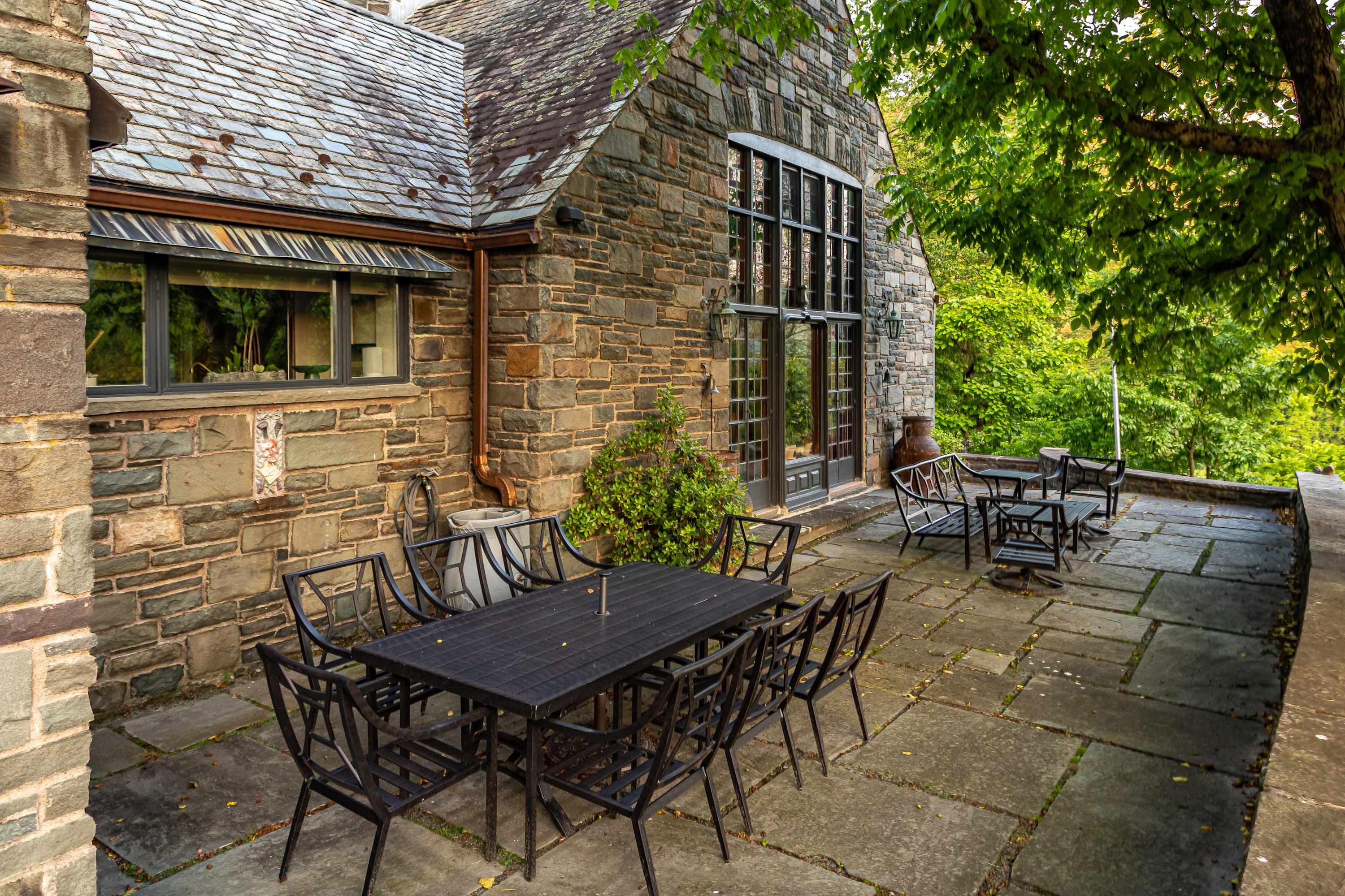 A spacious stone patio extends along the side of a charming stone house with a slate roof, featuring a large black outdoor dining table with eight chairs and additional seating areas, all nestled amidst vibrant green trees.