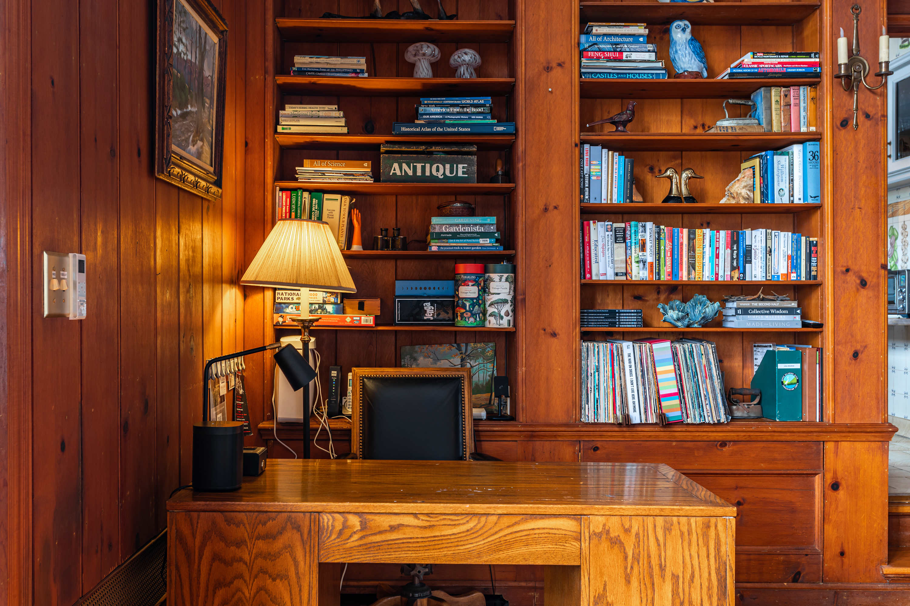 A dedicated office nook within a wood-paneled room, featuring a wooden desk with a black office chair and a desk lamp. Built-in bookshelves filled with books and decor surround the workspace.