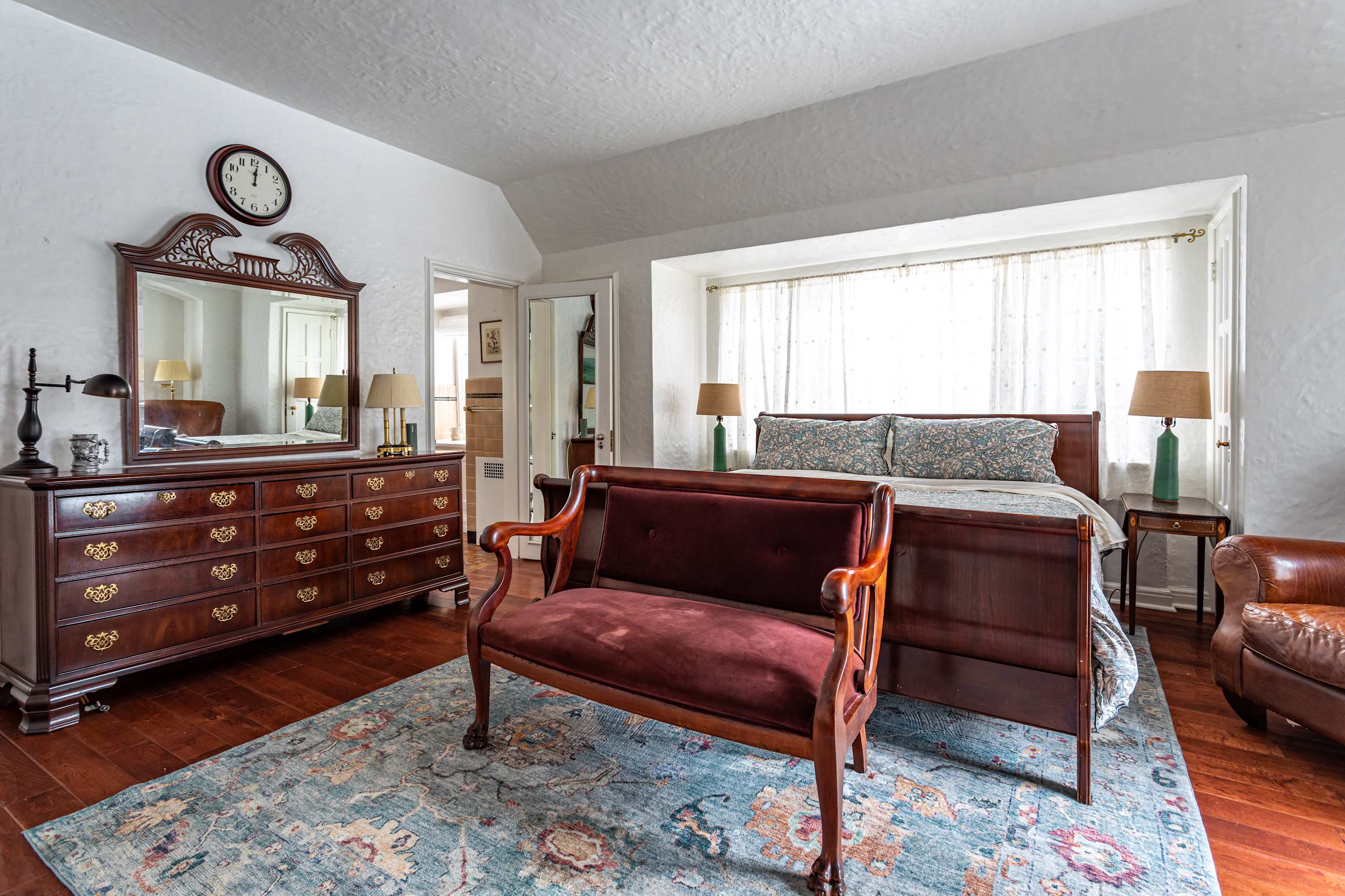 A wide view of a bedroom featuring a large wooden dresser with a mirror, a velvet upholstered bench, and a large bed with patterned bedding, all on dark hardwood floors with an area rug.