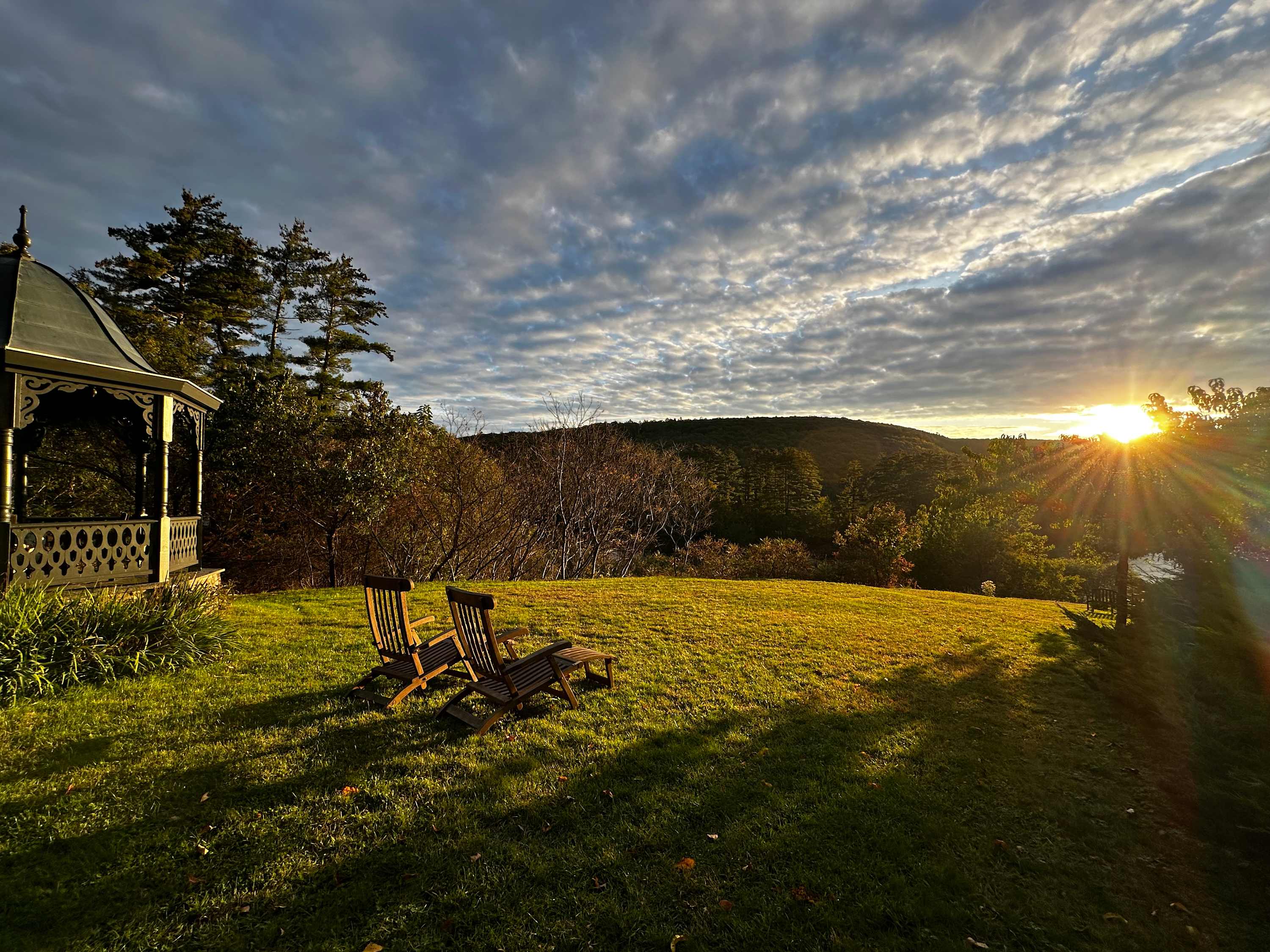 A picturesque sunset illuminates a grassy lawn with two wooden Adirondack chairs, positioned near a dark ornate gazebo, all overlooking a distant tree-covered mountain range under a dramatic sky.