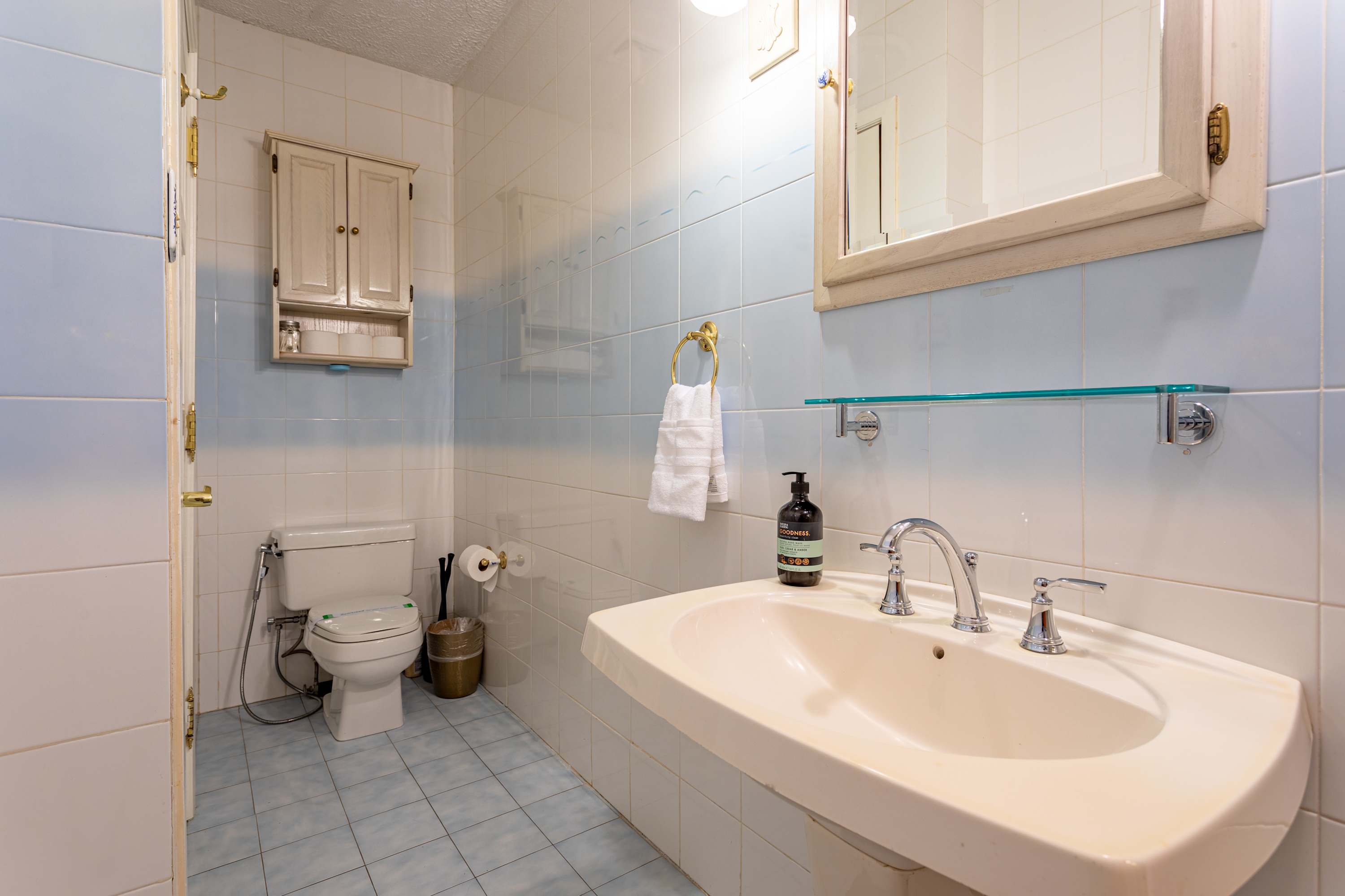 Full bathroom with light blue and white tiled walls, featuring a toilet with a bidet attachment and a white pedestal sink with chrome fixtures. A small medicine cabinet is mounted above the toilet for storage.