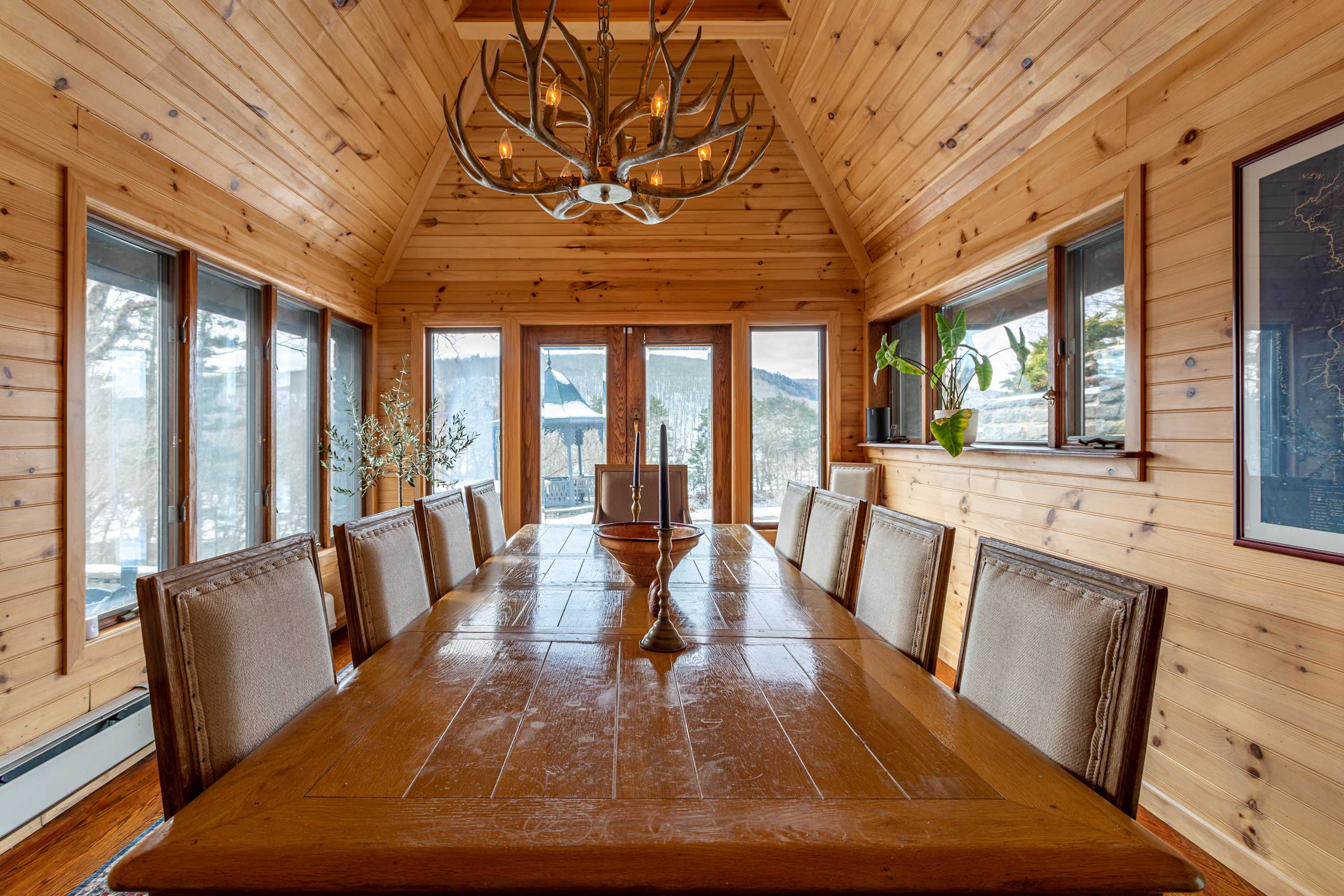 Expansive dining room with natural wood paneling on walls and a vaulted ceiling, featuring a long wooden dining table set for many guests. Large windows offer panoramic views of a snowy landscape, bringing the outdoors in.