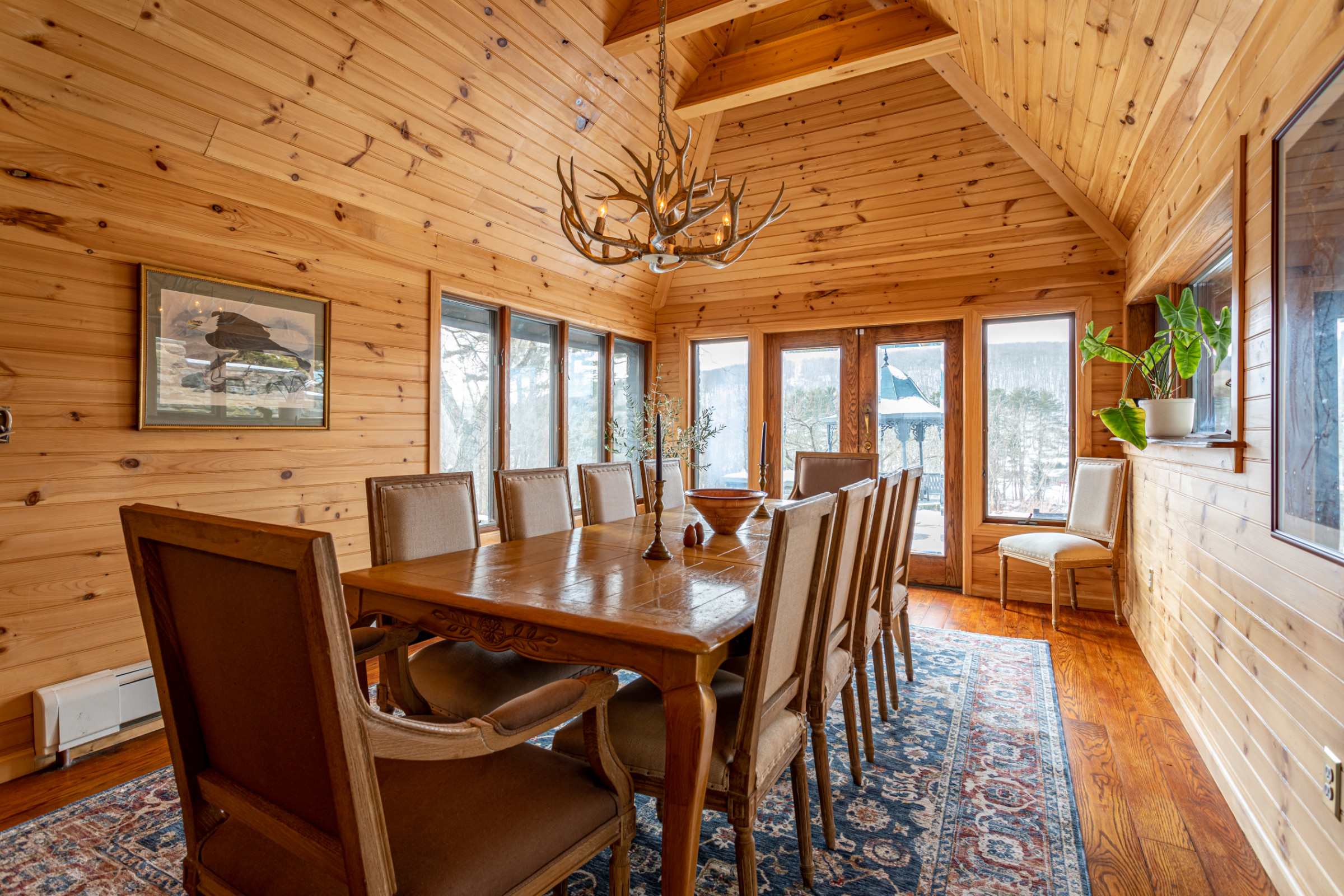 A spacious dining room features light wood-paneled walls and a vaulted ceiling, with a long wooden dining table surrounded by ten upholstered chairs. Large windows and glass doors offer panoramic views of the snowy landscape.