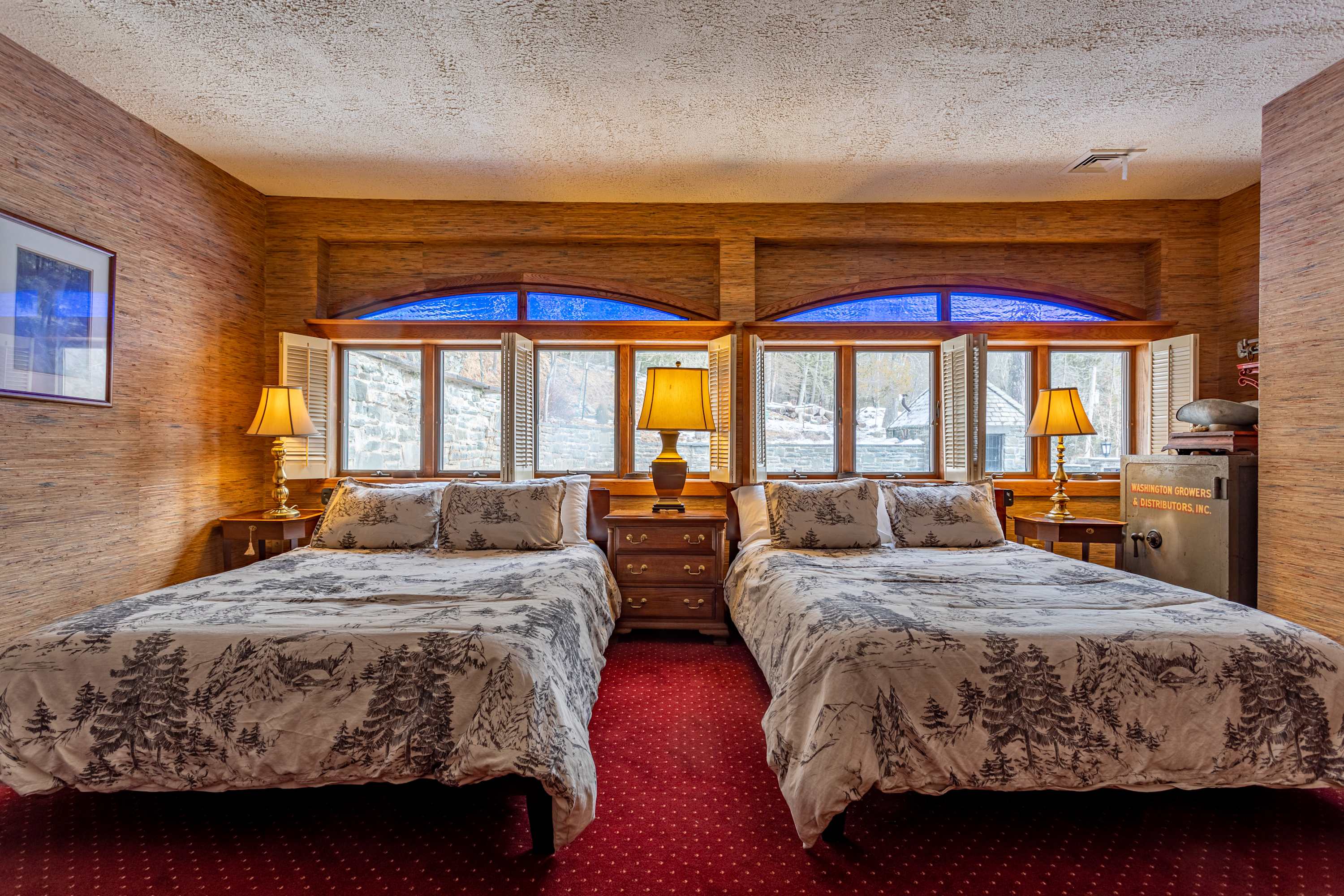 Another view of the bedroom showing two queen beds with patterned bedding, flanked by wooden nightstands and lamps. Windows with white shutters and decorative blue glass accents are visible above the beds, adding a unique touch.