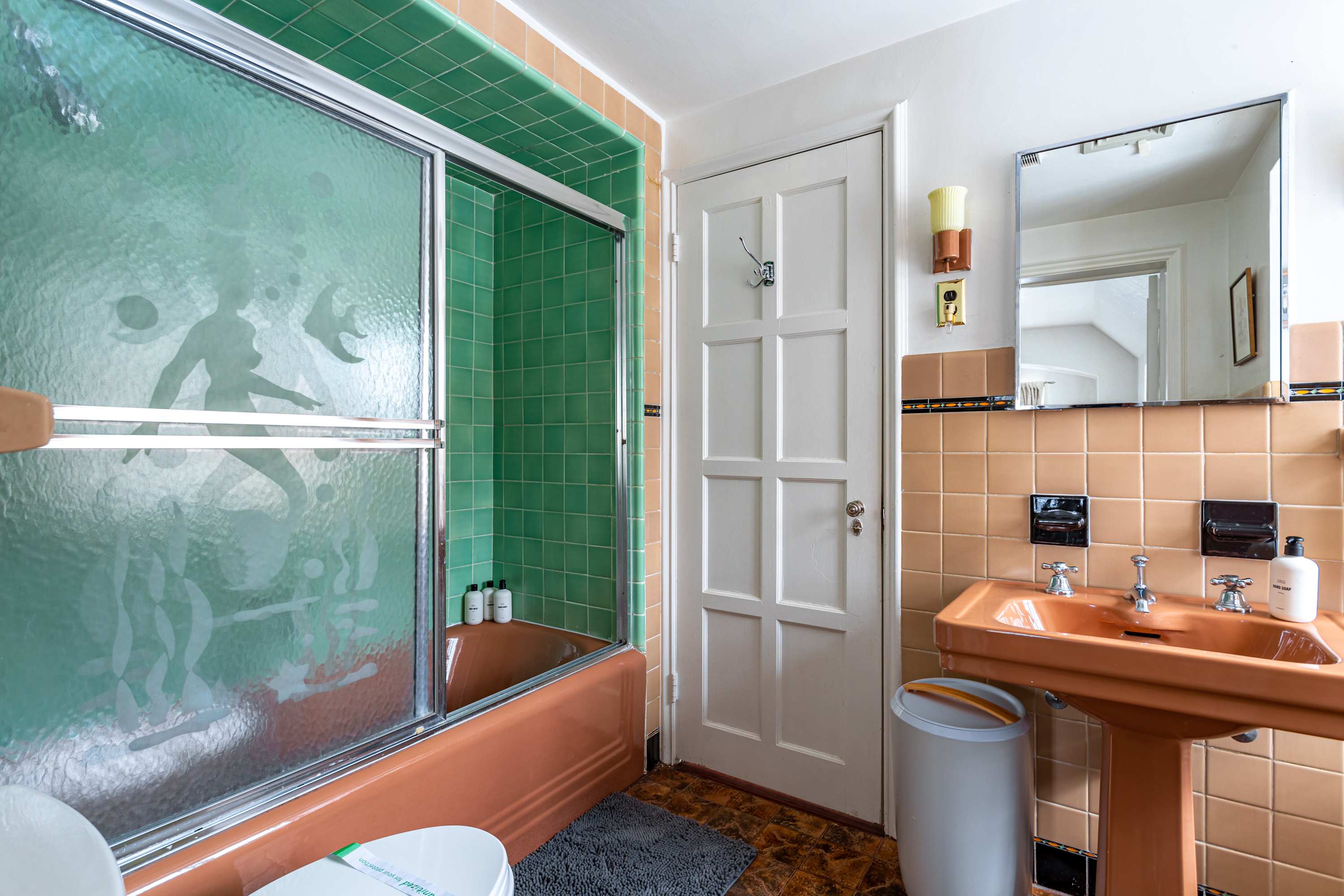 A retro-style bathroom with peach-colored pedestal sink and bathtub, green tiled walls in the shower area, and a glass shower door with etched designs.