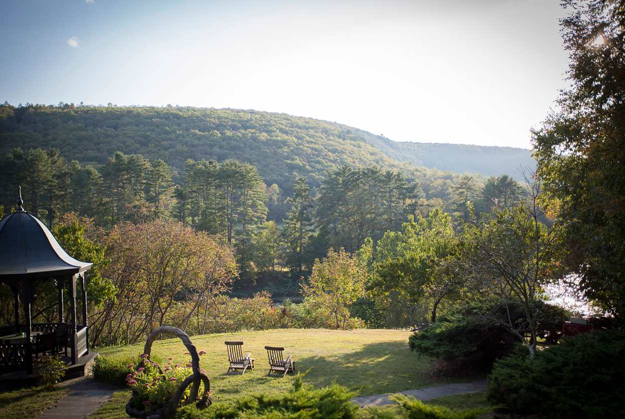 A lush green lawn features two wooden Adirondack chairs and an ornate dark gazebo, offering a serene view of a vast, tree-covered mountain landscape under a bright sky.