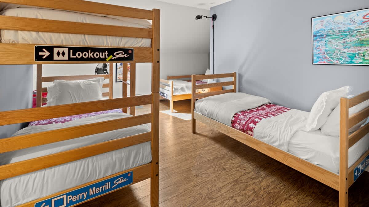 A spacious group bedroom with light gray walls and wooden floors, furnished with two wooden bunk beds and two single beds, all with white bedding and red patterned blankets. Ski-themed decor is visible on the walls.
