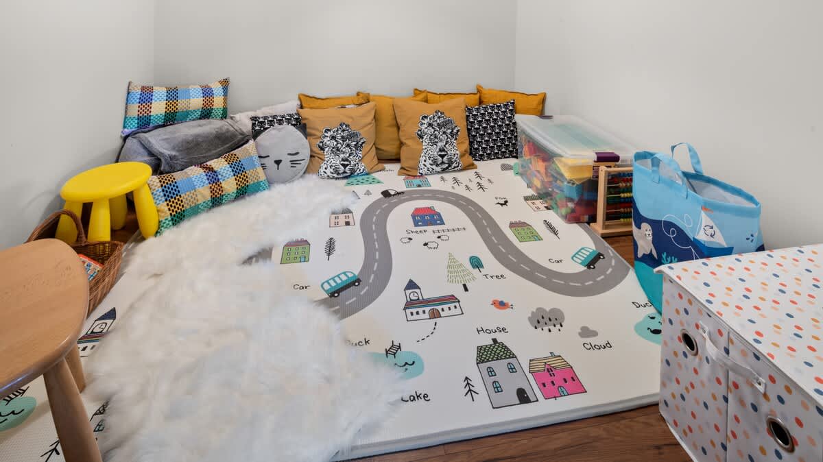 A vibrant children's play area with a road-themed mat on a wooden floor, surrounded by colorful pillows and storage bins filled with toys. A small yellow stool and a white furry rug enhance the playful and inviting atmosphere.