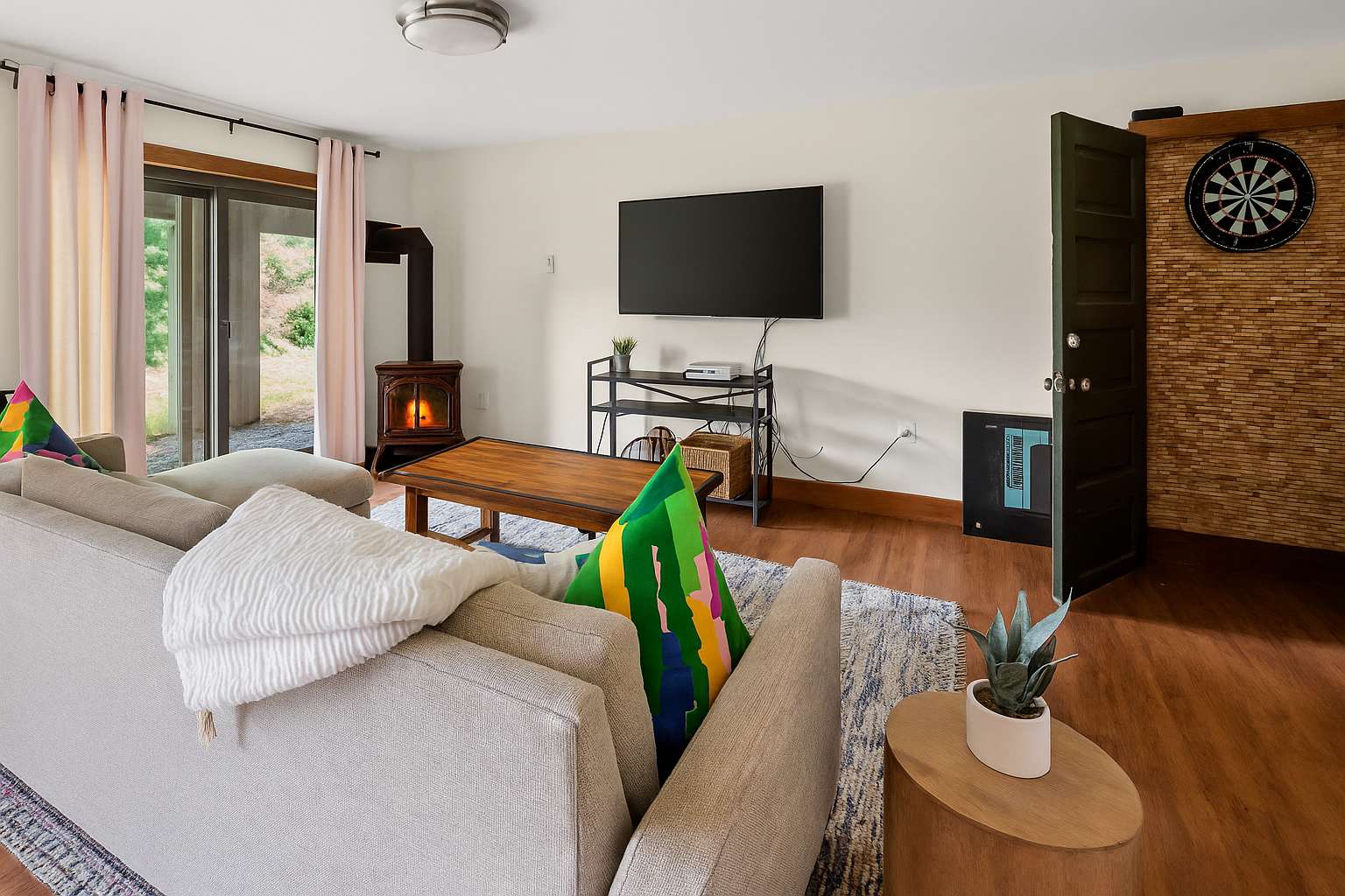 A cozy living room featuring light hardwood floors, a comfortable sectional sofa with colorful pillows, a wall-mounted TV, and a rustic wood-burning stove in the corner.