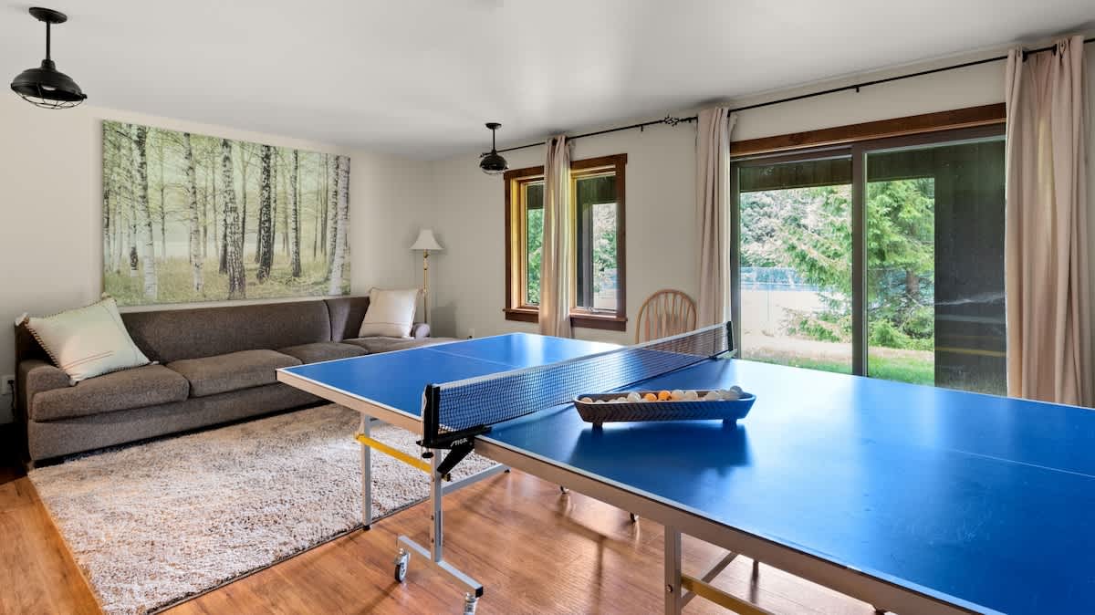 A bright game room with light hardwood floors, a blue ping-pong table in the foreground, and a comfortable sofa with a large nature-themed artwork on the wall.